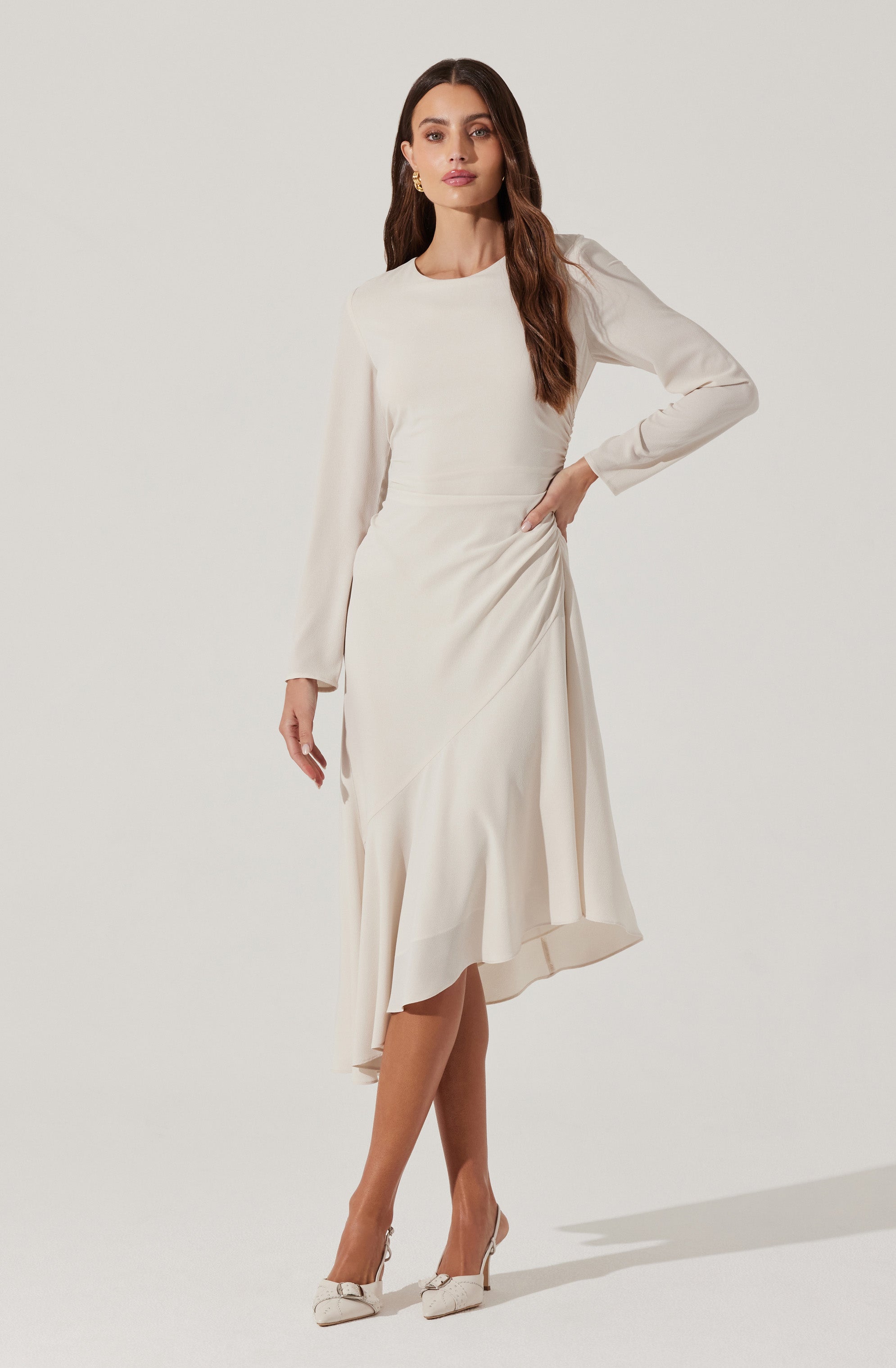 The woman is wearing a long, elegant, cream-colored dress with long sleeves and a round neckline. The dress features a fitted waist and a asymmetrical, flowy hemline that falls below the knees. She is also wearing light-colored high heels with a bow detail on the front.