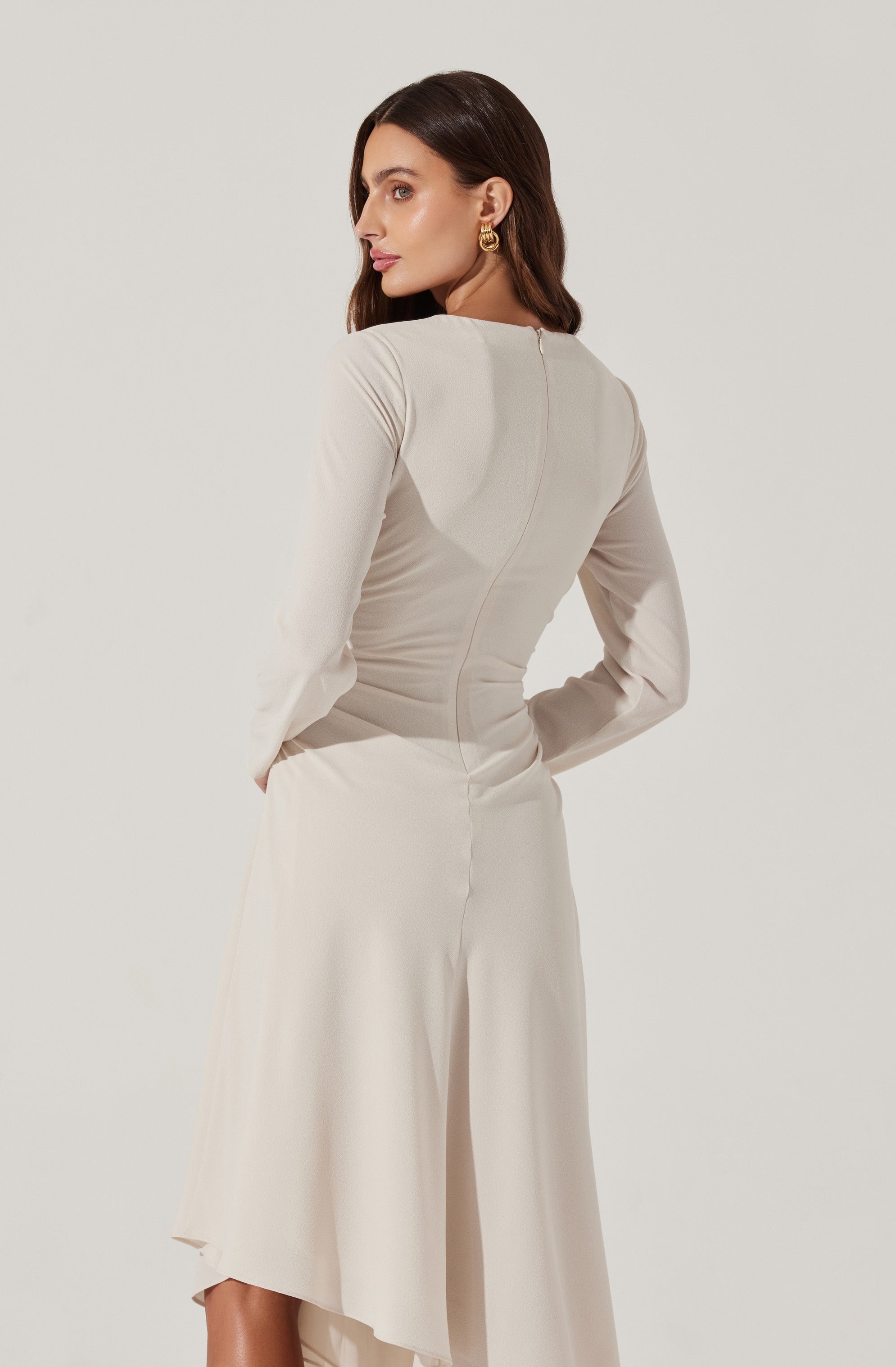 A woman is wearing a long-sleeved, cream-colored dress with a fitted bodice and a flowy, asymmetrical skirt. The dress has a visible seam that runs vertically down the back and appears to be made of a smooth, satin-like fabric. She is accessorizing with gold hoop earrings.