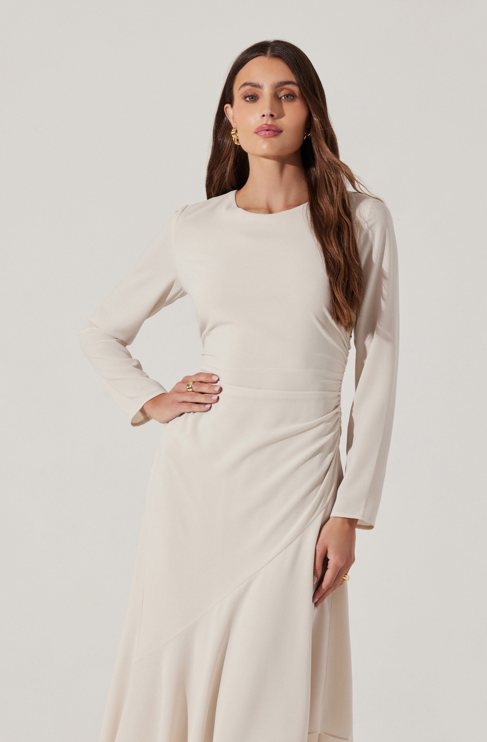 A woman is wearing a long, elegant, cream-colored dress with a high neckline and long sleeves. The dress features a smooth, satin-like fabric with subtle draping details along the waist and skirt. She accessorizes with gold earrings and rings.