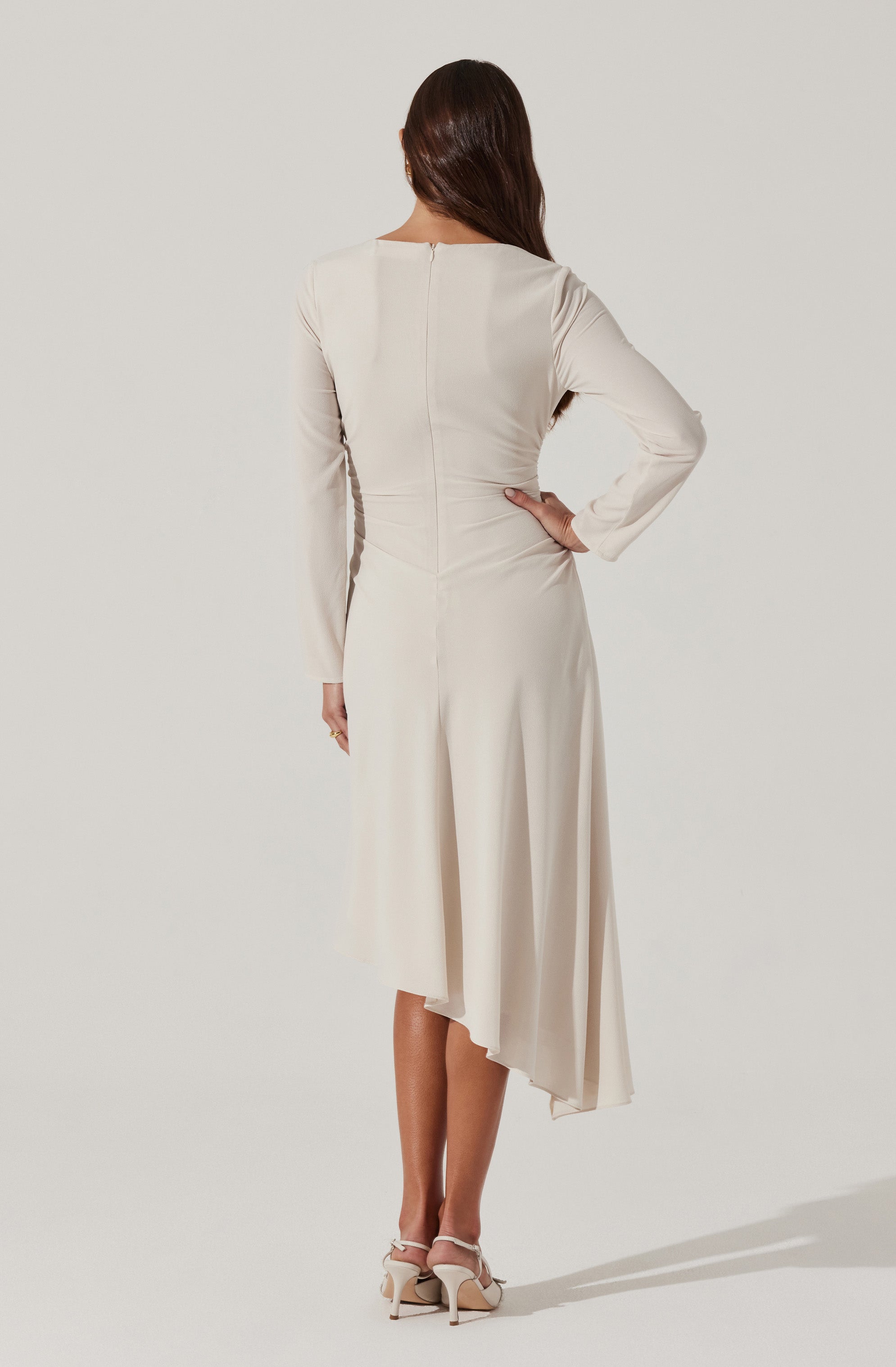 A woman is wearing a long, elegant dress with a fitted bodice and long sleeves. The dress is made of a smooth, light-colored fabric and features a zipper closure running down the back. The skirt has an asymmetrical hemline, which is shorter in the front and longer in the back, creating a flowing, layered effect. She is also wearing a pair of light-colored high-heeled shoes with ankle straps.