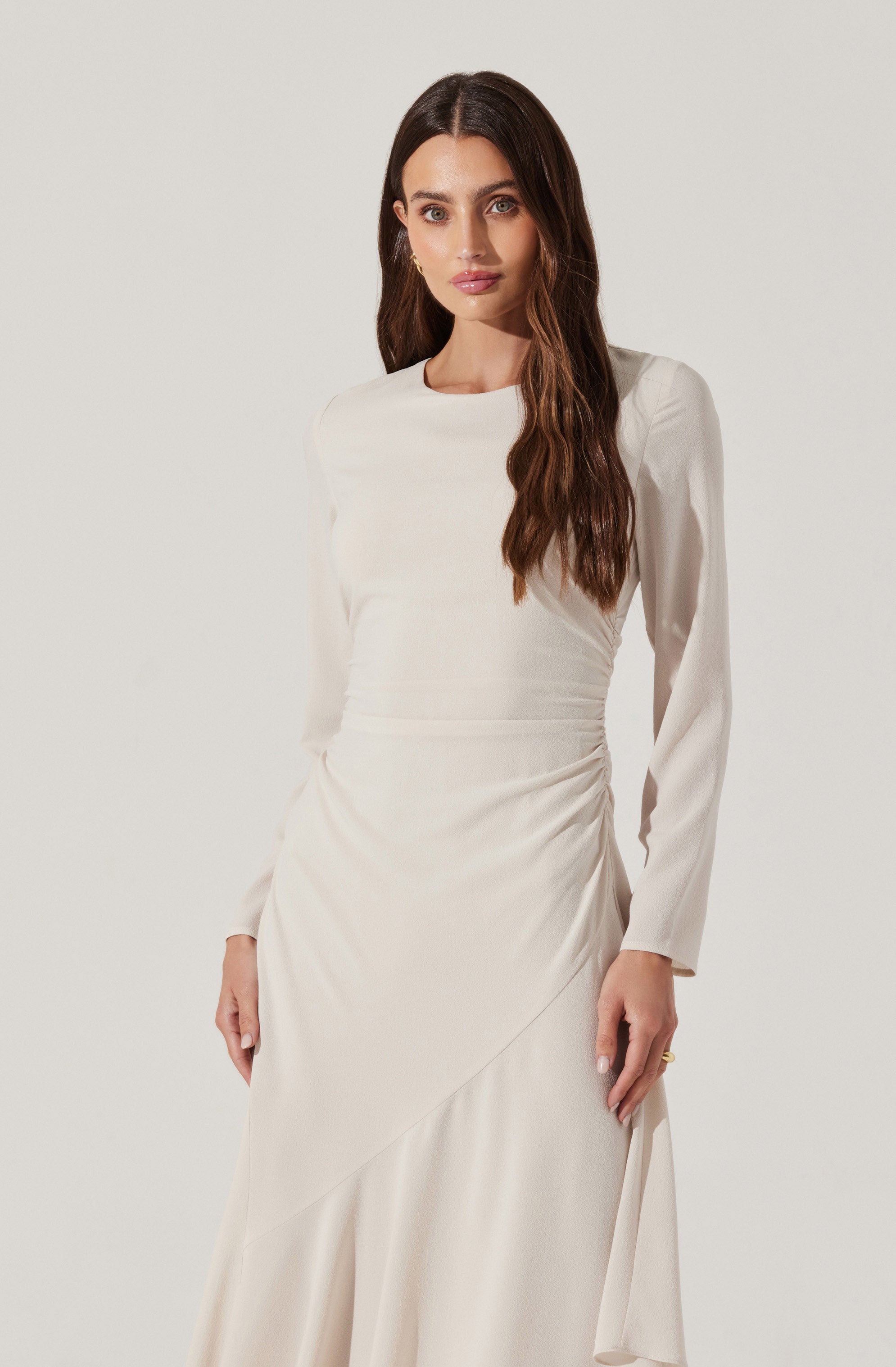 A woman is wearing a long, elegant, cream-colored dress. The dress has long sleeves that are slightly puffed at the shoulders, a high, round neckline, and a fitted waist. The fabric appears smooth and satin-like, with subtle draping and gathering details on the skirt, which has a slight asymmetrical design.