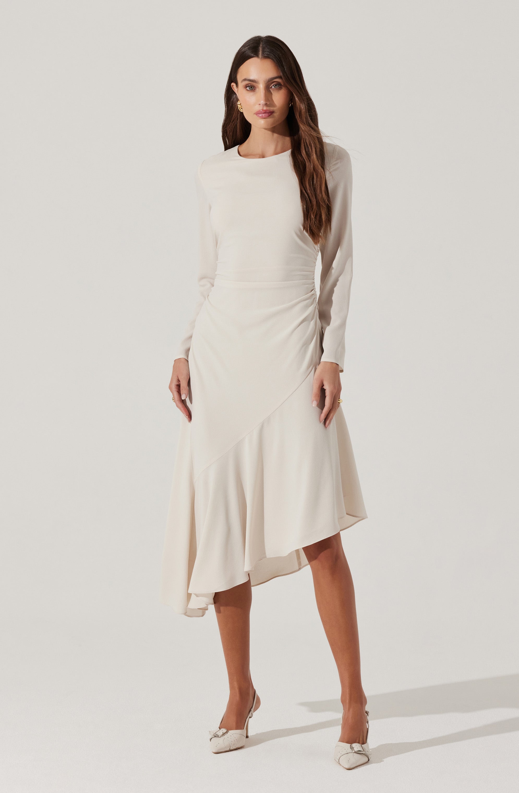 A woman is wearing a long, elegant, cream-colored dress with long sleeves. The dress has a fitted bodice and features an asymmetrical, flowing skirt that falls mid-calf. She completes her look with a pair of light-colored heels.