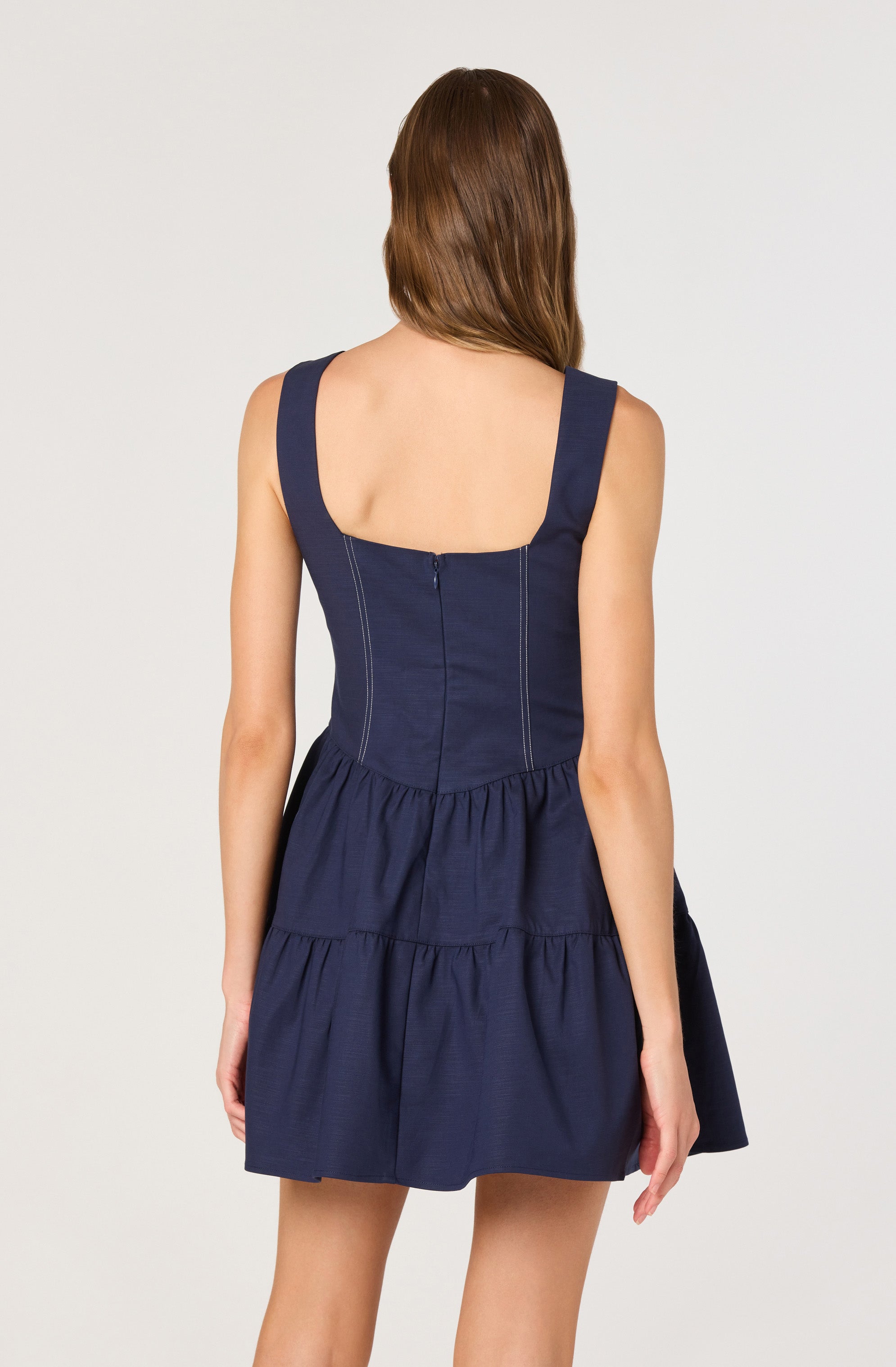 A woman is wearing a sleeveless navy blue dress with wide shoulder straps. The dress has a fitted bodice with visible vertical seam lines and a back zipper closure. The skirt portion of the dress is gathered with a tiered design, creating a full and ruffled appearance.