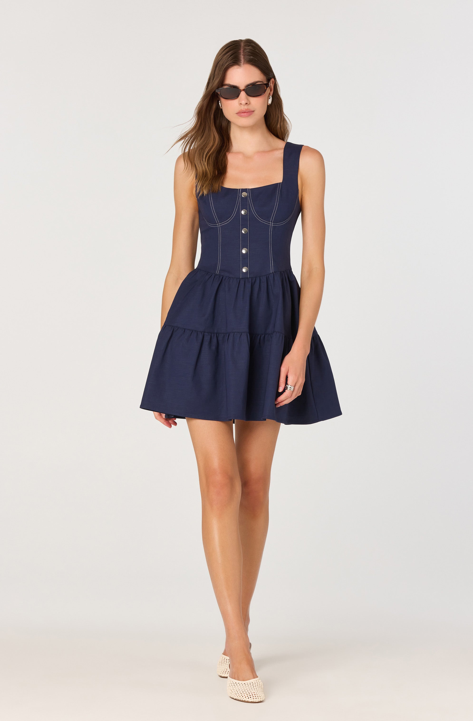 A woman is wearing a sleeveless, navy blue dress with wide shoulder straps. The fitted bodice features decorative white stitching and a row of buttons down the front. The skirt part is gathered at the waist, creating a full, layered, and slightly flared silhouette. She accessorizes with dark sunglasses and light-colored woven shoes.
