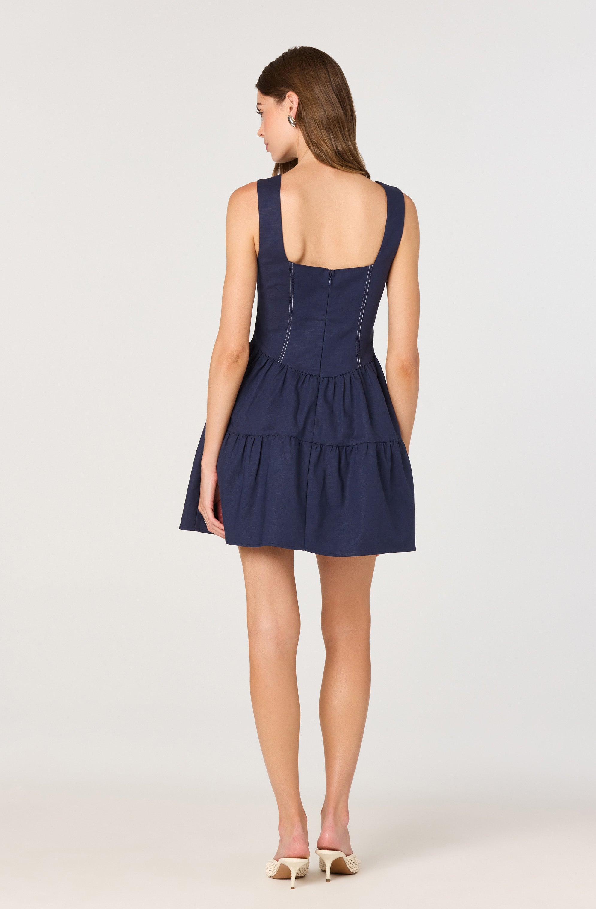 A woman is wearing a sleeveless navy blue dress with wide shoulder straps. The dress features a fitted bodice with vertical seams and a square neckline at the back. The skirt has a gathered, tiered design, creating a ruffled effect. The woman is also wearing light-colored, open-toed high-heeled shoes.