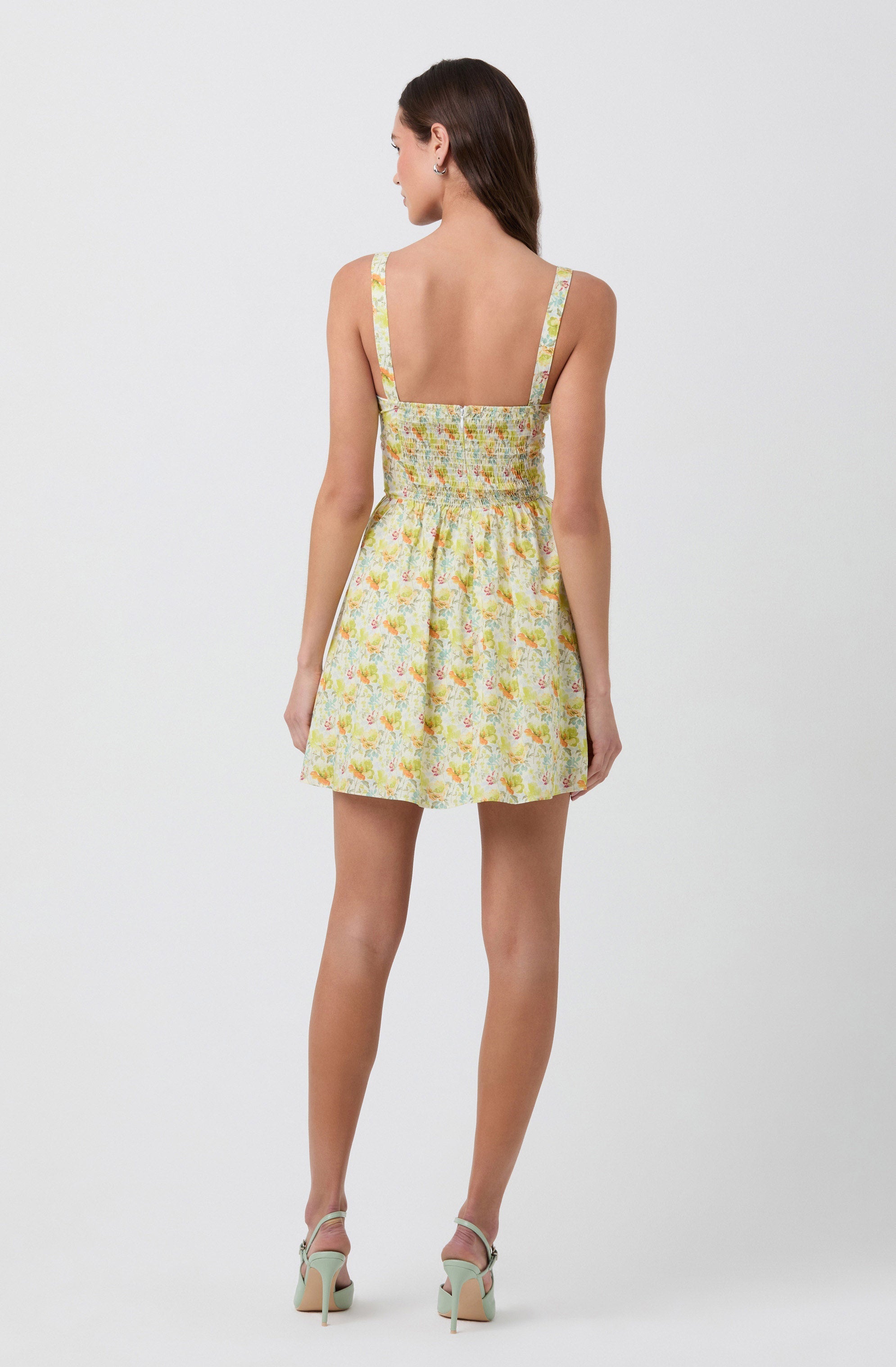 A woman is wearing a sleeveless dress with wide straps. The dress features a pastel floral pattern in shades of yellow, orange, and green. It has a fitted, smocked bodice and a high waistline, with the skirt flaring out slightly to a short length above the knees. She is also wearing high-heeled sandals in a light color.