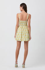 A woman is wearing a sleeveless dress with wide straps. The dress features a pastel floral pattern in shades of yellow, orange, and green. It has a fitted, smocked bodice and a high waistline, with the skirt flaring out slightly to a short length above the knees. She is also wearing high-heeled sandals in a light color.