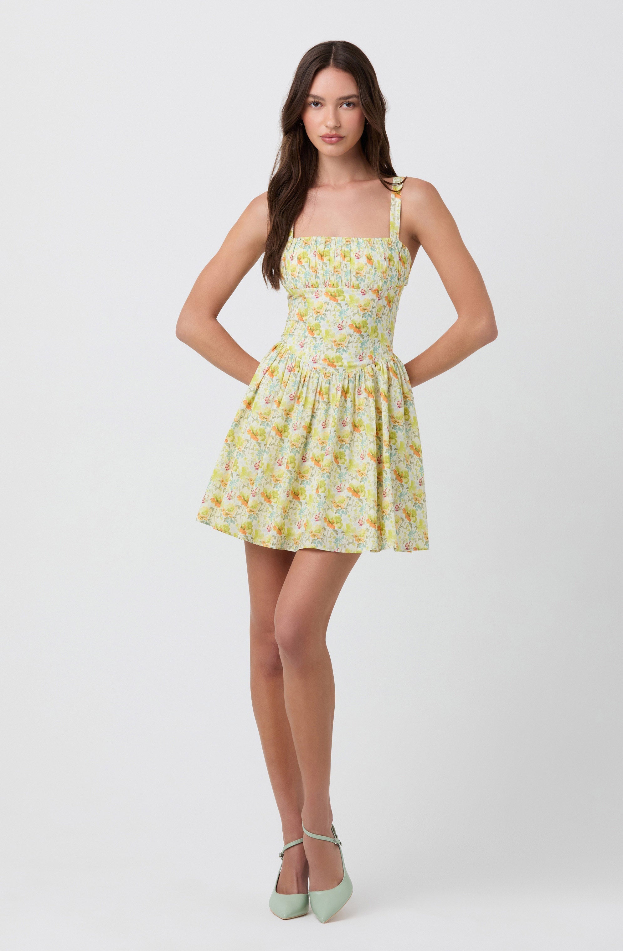 A young woman wearing a sleeveless, fitted dress with a square neckline and wide shoulder straps. The dress features a floral pattern in pastel colors and has a gathered, flared skirt that falls above the knees. She is also wearing light-colored high heels.