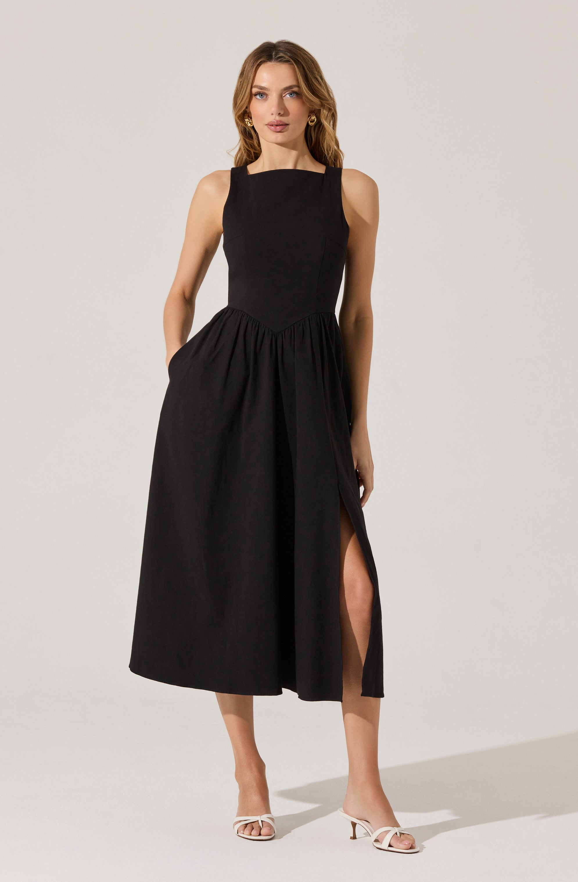 A woman is wearing a sleeveless, black midi dress with a fitted bodice and a gathered skirt that features a thigh-high slit on the left side. She is also wearing white, open-toe high-heeled sandals and gold hoop earrings.
