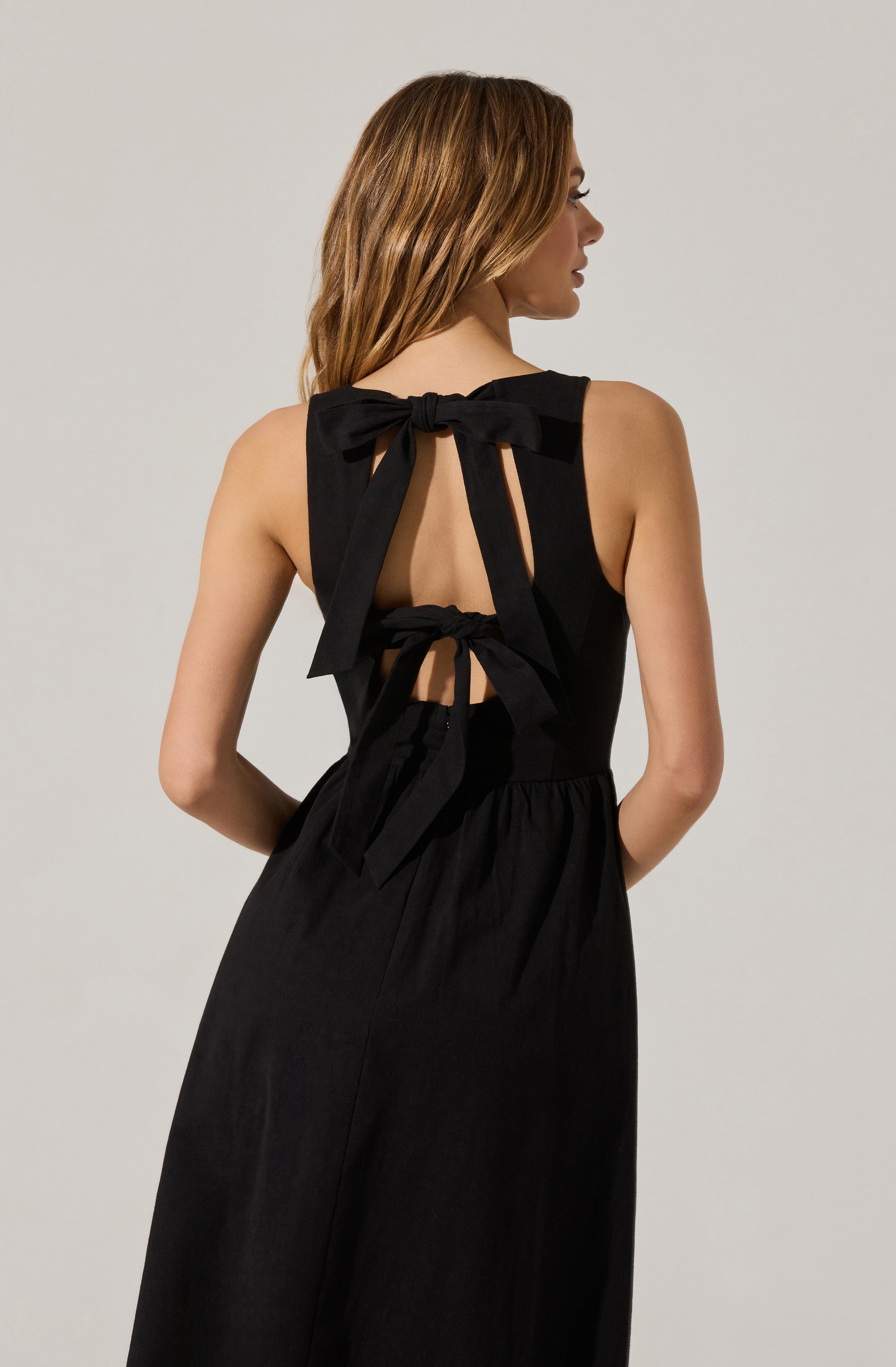 A woman is wearing a sleeveless black dress with large bows on the back. The dress has open back details with fabric ties that create a bow at the neckline and a bow at the mid-back. The fabric appears to be lightweight and flowy.