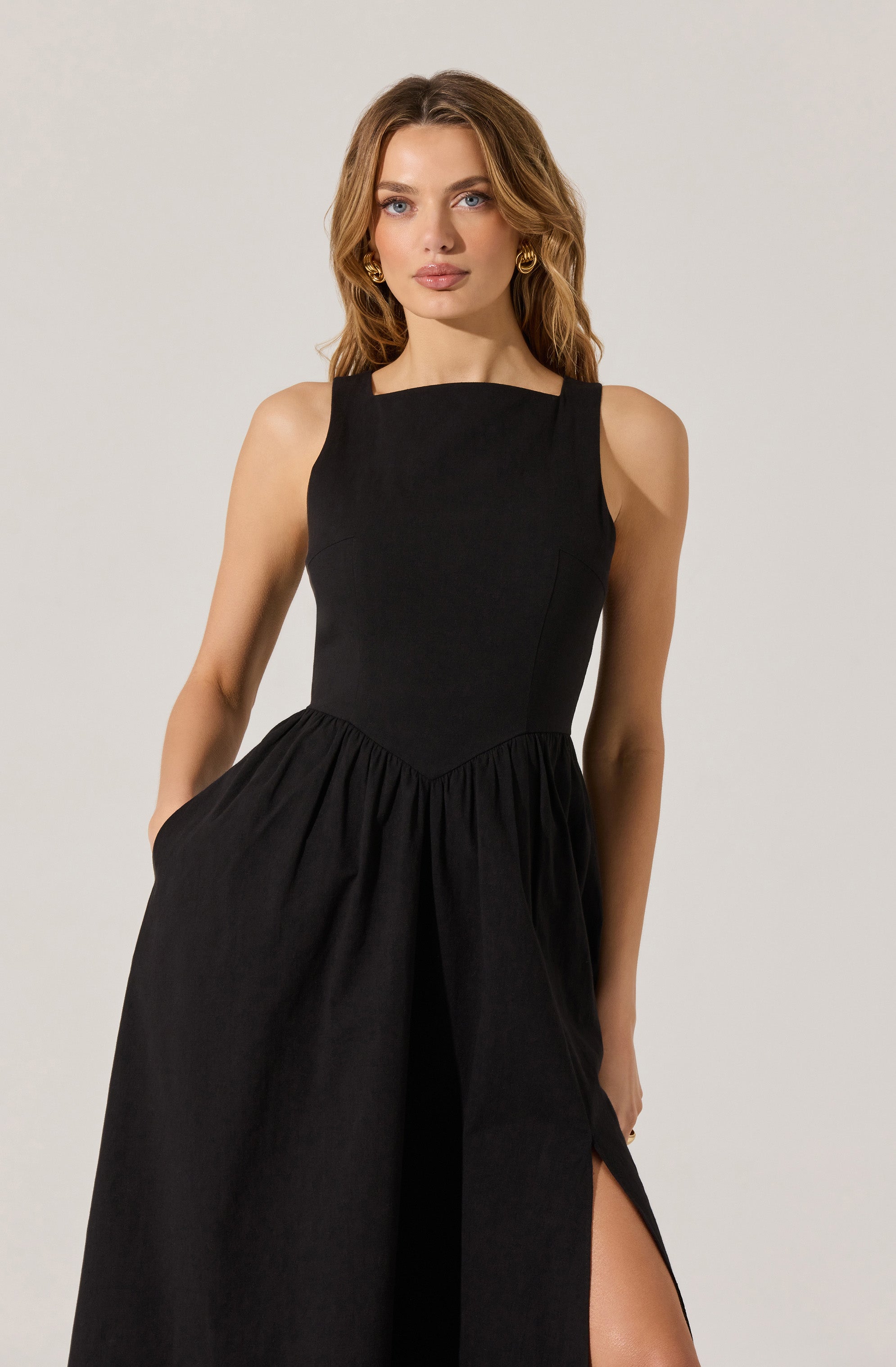 A woman is wearing a sleeveless black dress with a fitted bodice and wide shoulder straps. The skirt of the dress is gathered at the waist and has a side slit.