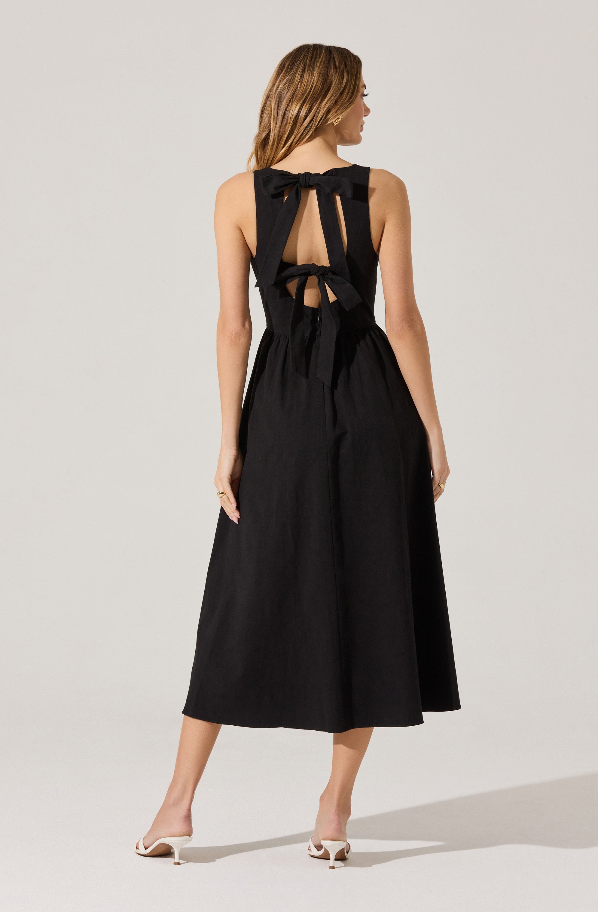 A woman is wearing a black sleeveless midi dress with a fitted waist and a full A-line skirt. The dress features straps tied into bows on her shoulders and back, with open back detailing. She pairs the dress with white high-heeled shoes.