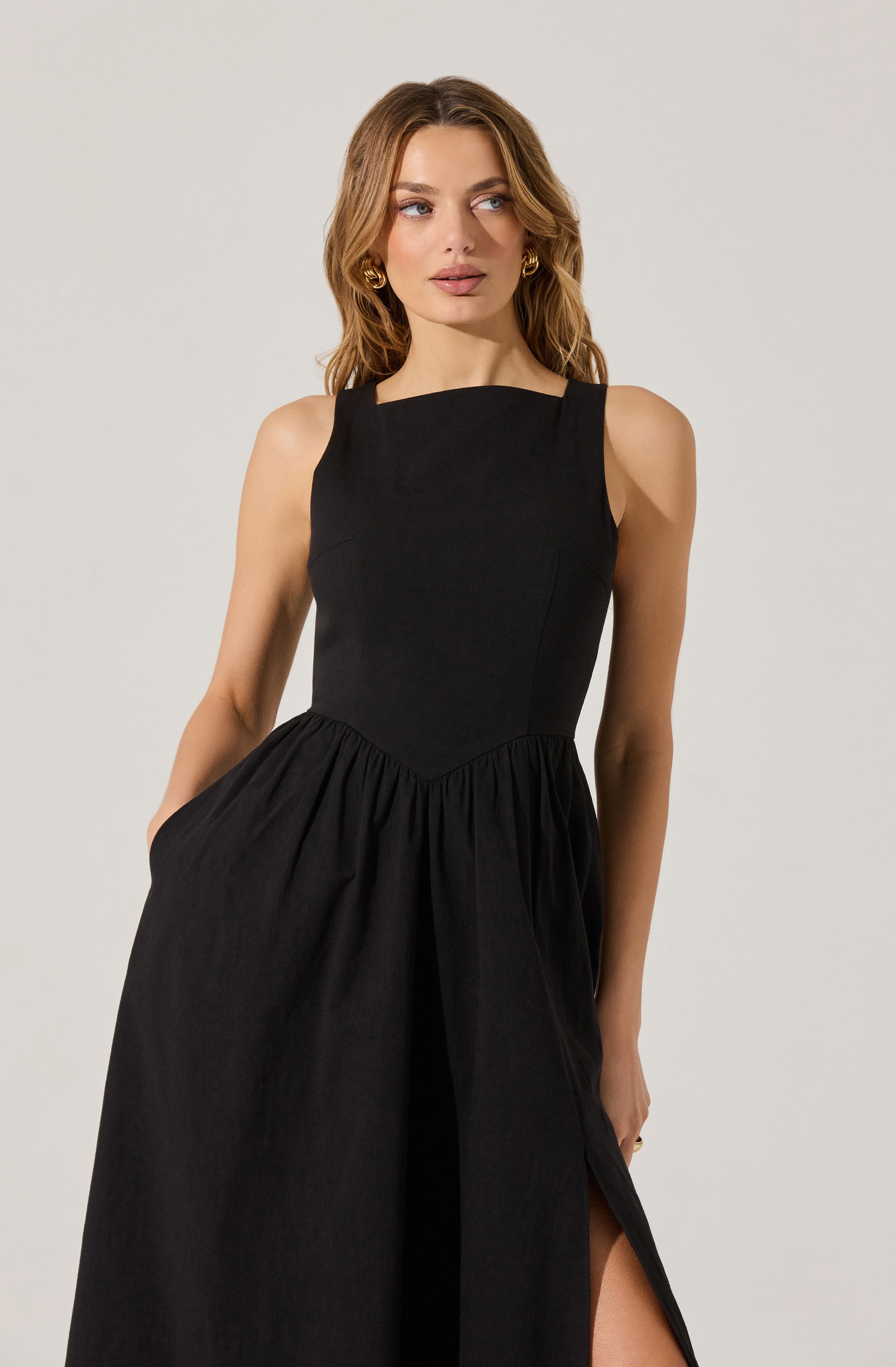 A woman is wearing a sleeveless black dress with wide straps that sit on her shoulders. The dress has a fitted bodice and a gathered, flowing skirt with a slit on the left side. She has wavy, shoulder-length hair and is accessorized with gold hoop earrings.