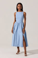 A woman is wearing a sleeveless, light blue dress with a fitted bodice and a full, gathered skirt that reaches mid-calf length. The dress features a high slit on one side, revealing her leg. She is also wearing white high-heeled sandals.