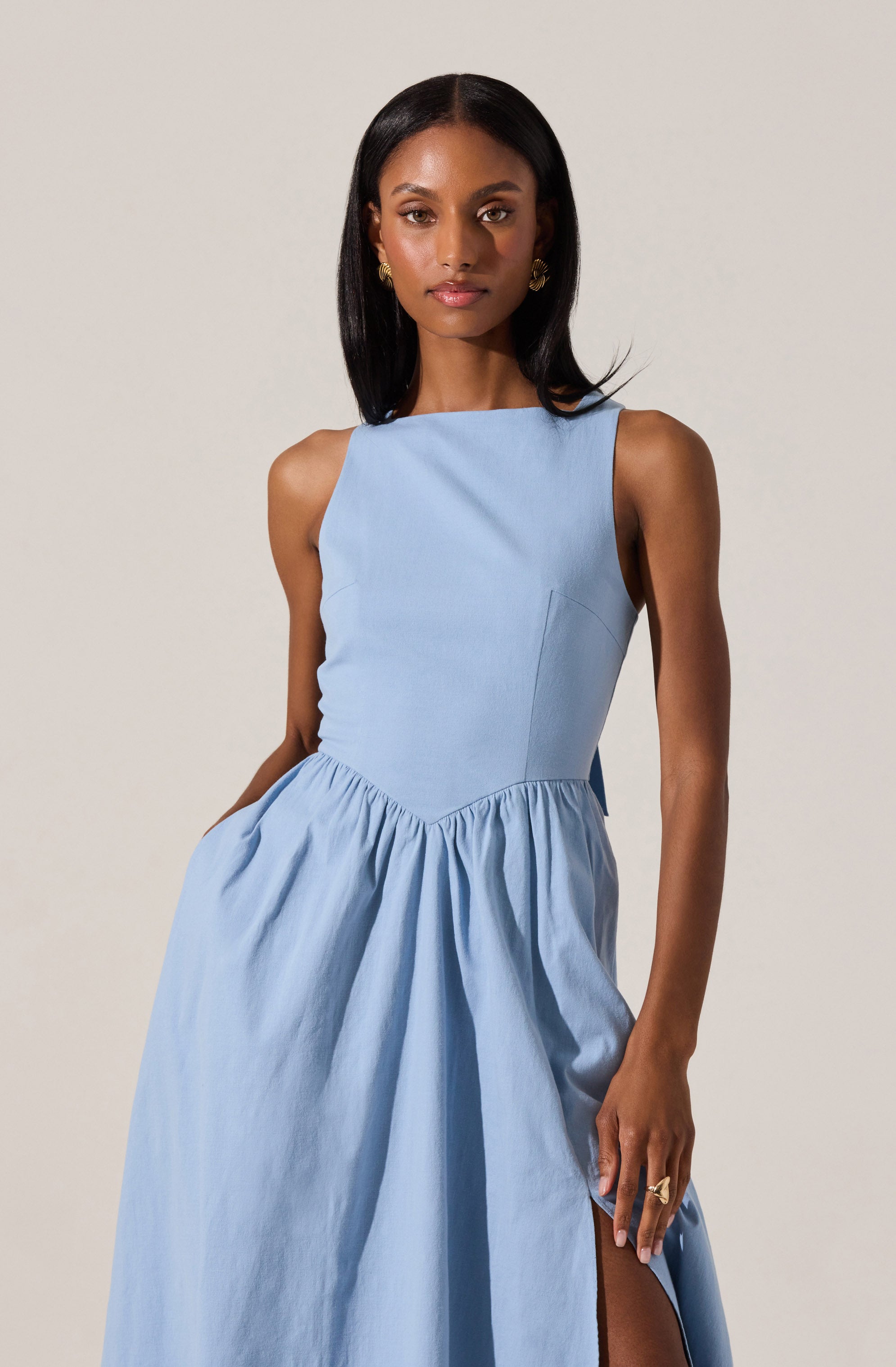 A woman is wearing a sleeveless, light blue dress with a fitted bodice and a high, rounded neckline. The dress features a gathered, full skirt with a side slit. She is accessorized with gold earrings and a ring.