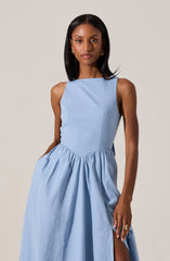 A woman is wearing a sleeveless, light blue dress with a fitted bodice and a high, rounded neckline. The dress features a gathered, full skirt with a side slit. She is accessorized with gold earrings and a ring.