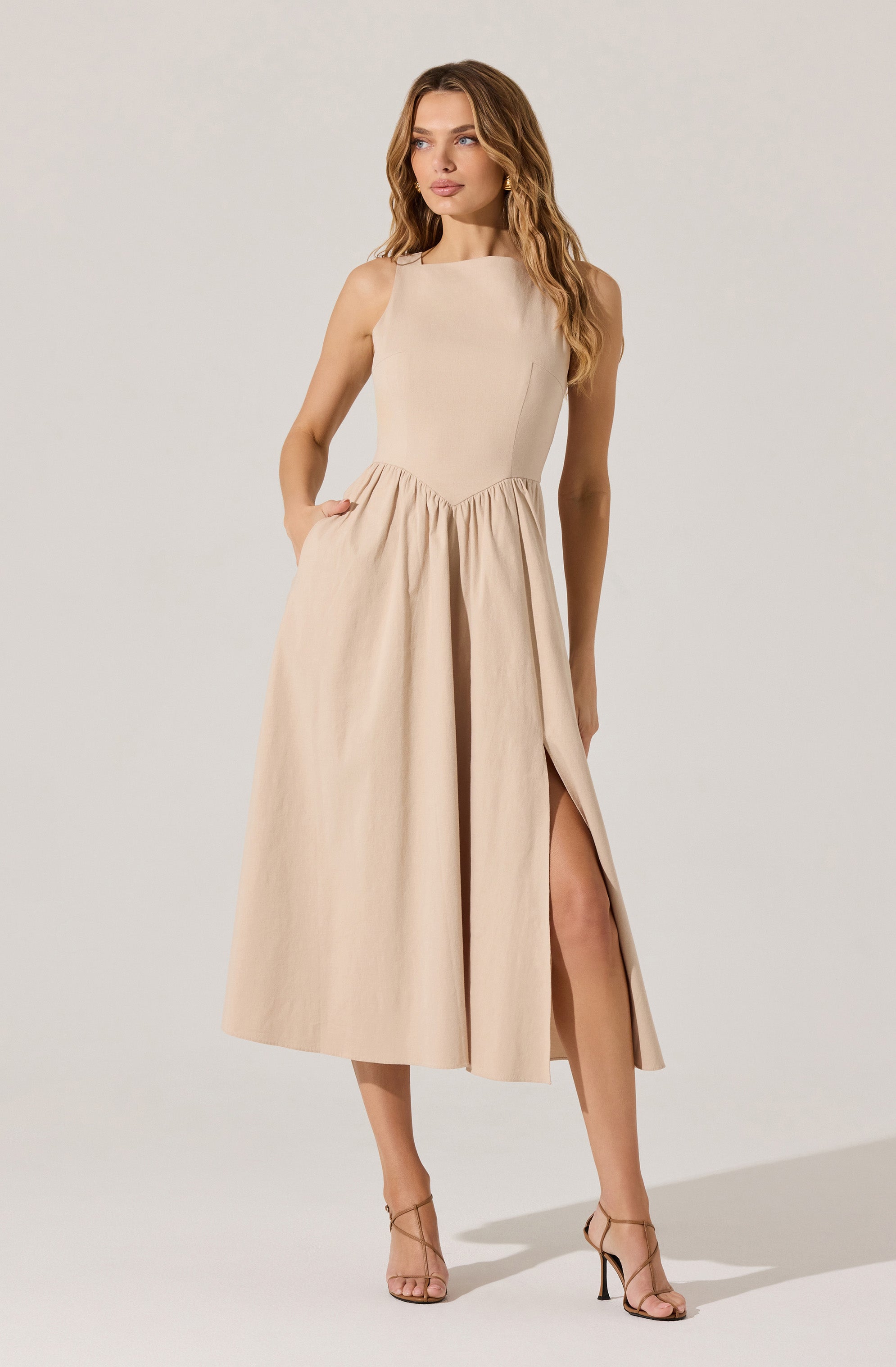 A woman is wearing a sleeveless, beige dress with a fitted bodice and a high, rounded neckline. The dress has a gathered waistline and a full, ankle-length skirt with a high slit on one side. She is also wearing nude, open-toe high-heeled sandals with thin straps.