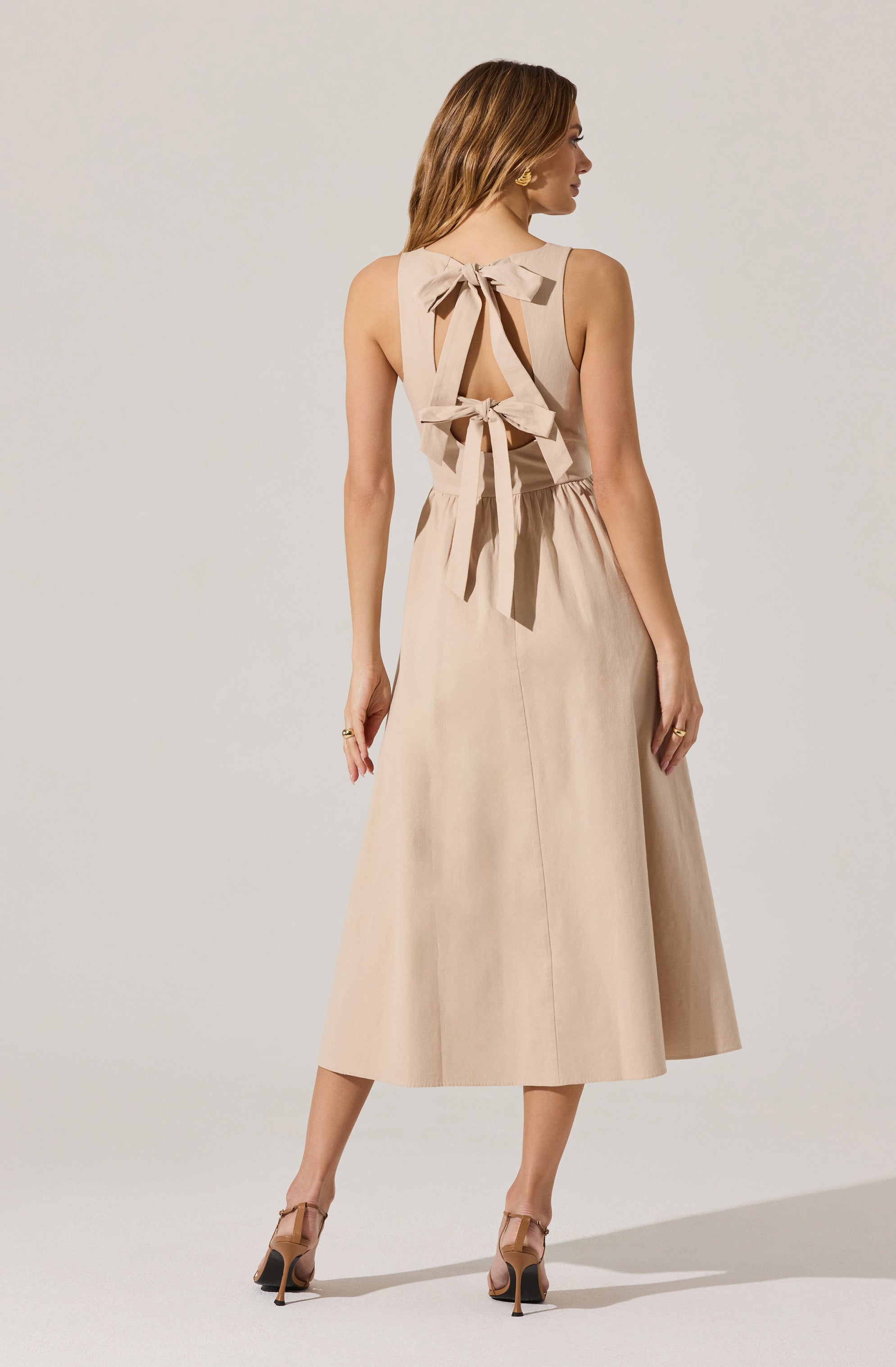 A woman is wearing a sleeveless, beige dress with a wide skirt that reaches mid-calf length. The dress features a large bow detail at the back with two bows tied at the shoulders and one at the waist, creating a decorative and open-back design. She is also wearing nude high-heeled sandals with ankle straps.