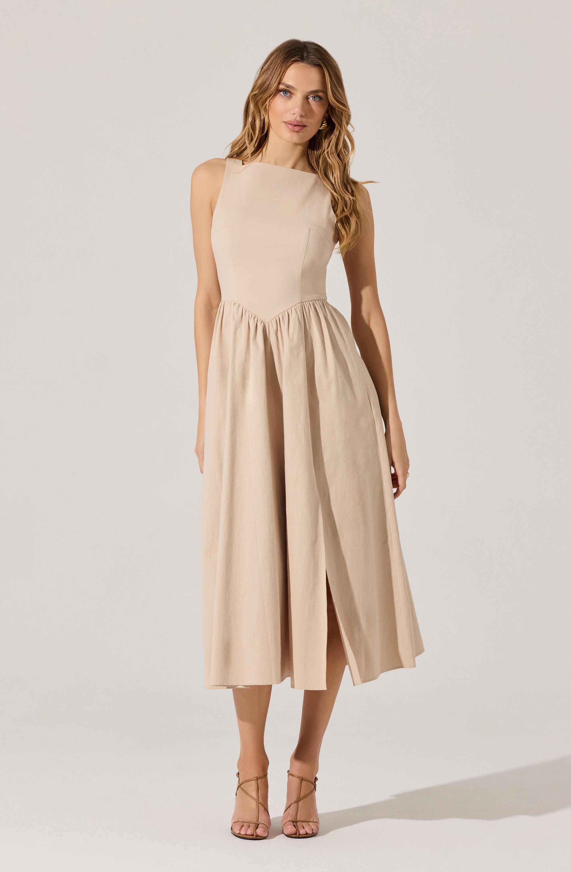 A woman is wearing a sleeveless, beige dress with a fitted bodice and a full, pleated skirt that falls below the knees. The dress has a high, rounded neckline and a slit in the front of the skirt. She is also wearing nude, strappy high-heeled sandals.