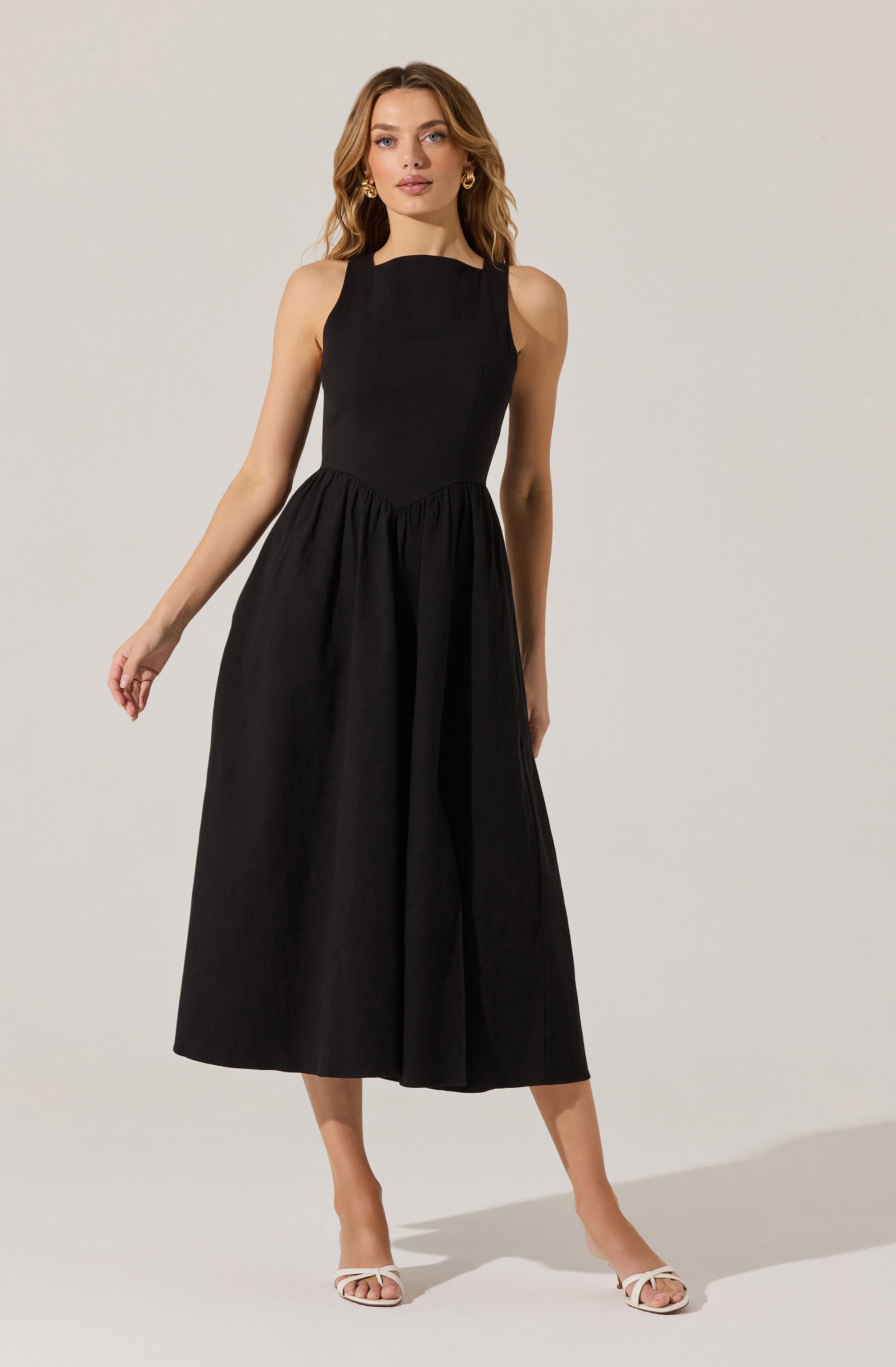 A woman is wearing a sleeveless, black, full-length dress with a fitted bodice and a gathered waist. She pairs the dress with white open-toe sandals and gold hoop earrings.