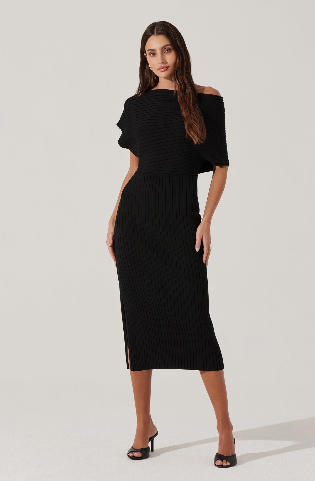 A woman is wearing a black knit outfit consisting of a loose off-the-shoulder top with wide sleeves and a fitted, high-waisted, ribbed midi skirt with a side slit. She pairs the outfit with black open-toe high heels.