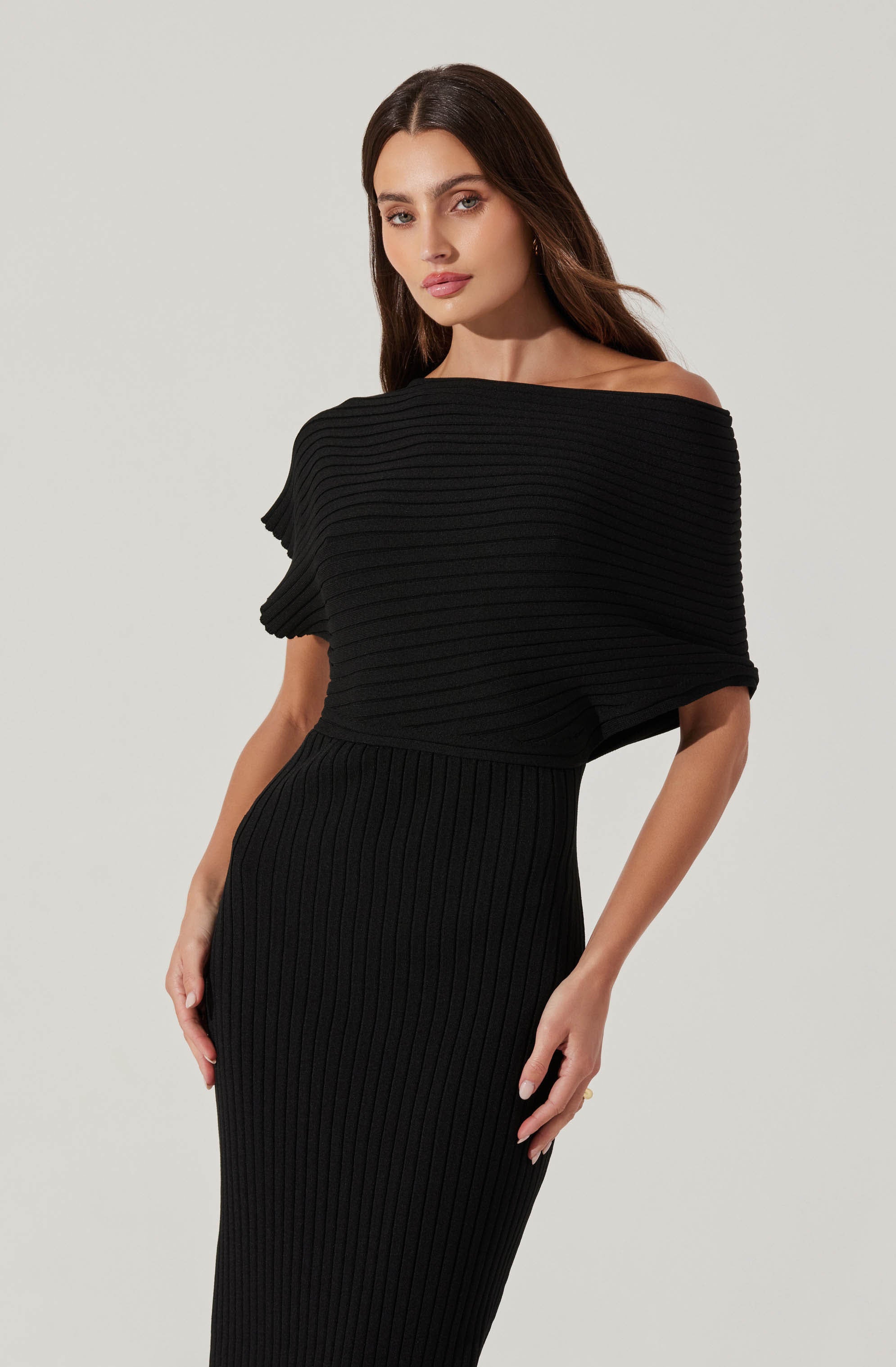 A woman is wearing a black, ribbed knit dress. The dress features a wide neckline that drapes off one shoulder, creating an off-the-shoulder look. The top part of the dress has horizontal ribbing, while the skirt has vertical ribbing, creating a contrast in the knit pattern. The dress is form-fitting and elegant.