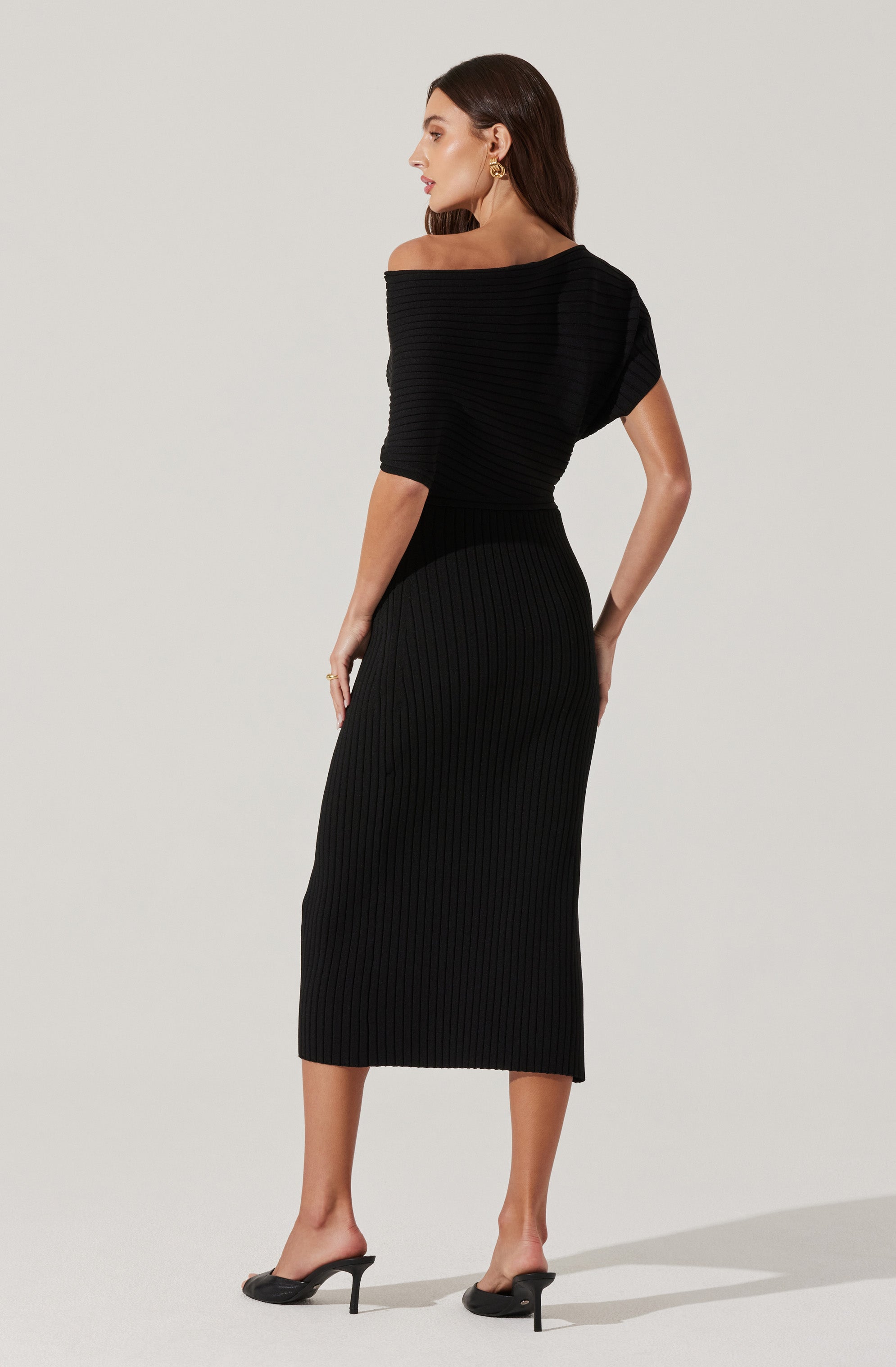 A woman is wearing a black, off-the-shoulder, ribbed knit dress that extends to mid-calf length. The dress features a horizontal ribbed pattern and short, loose sleeves. She is accessorized with gold hoop earrings and rings, and is wearing black open-toe high-heeled shoes.
