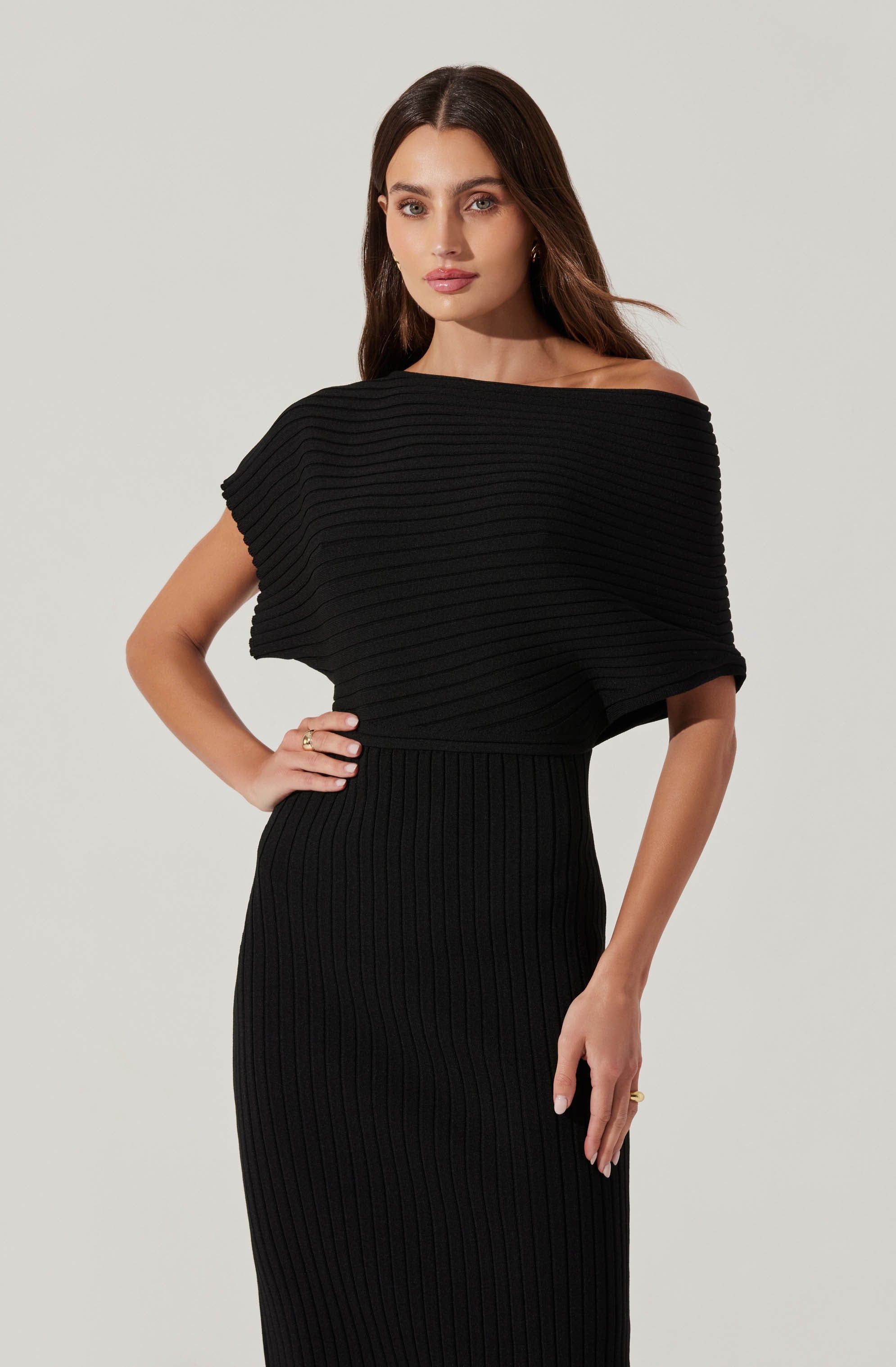 A woman is wearing a black, ribbed knit dress with a fitted skirt. The dress features an off-the-shoulder neckline and a loose, short-sleeved top section with horizontal ribbing. The skirt is high-waisted and extends to below the knees, with vertical ribbing throughout.