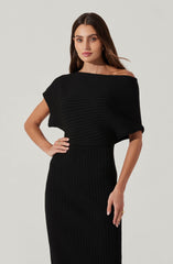 A woman is wearing a black, ribbed knit dress with a fitted skirt. The dress features an off-the-shoulder neckline and a loose, short-sleeved top section with horizontal ribbing. The skirt is high-waisted and extends to below the knees, with vertical ribbing throughout.