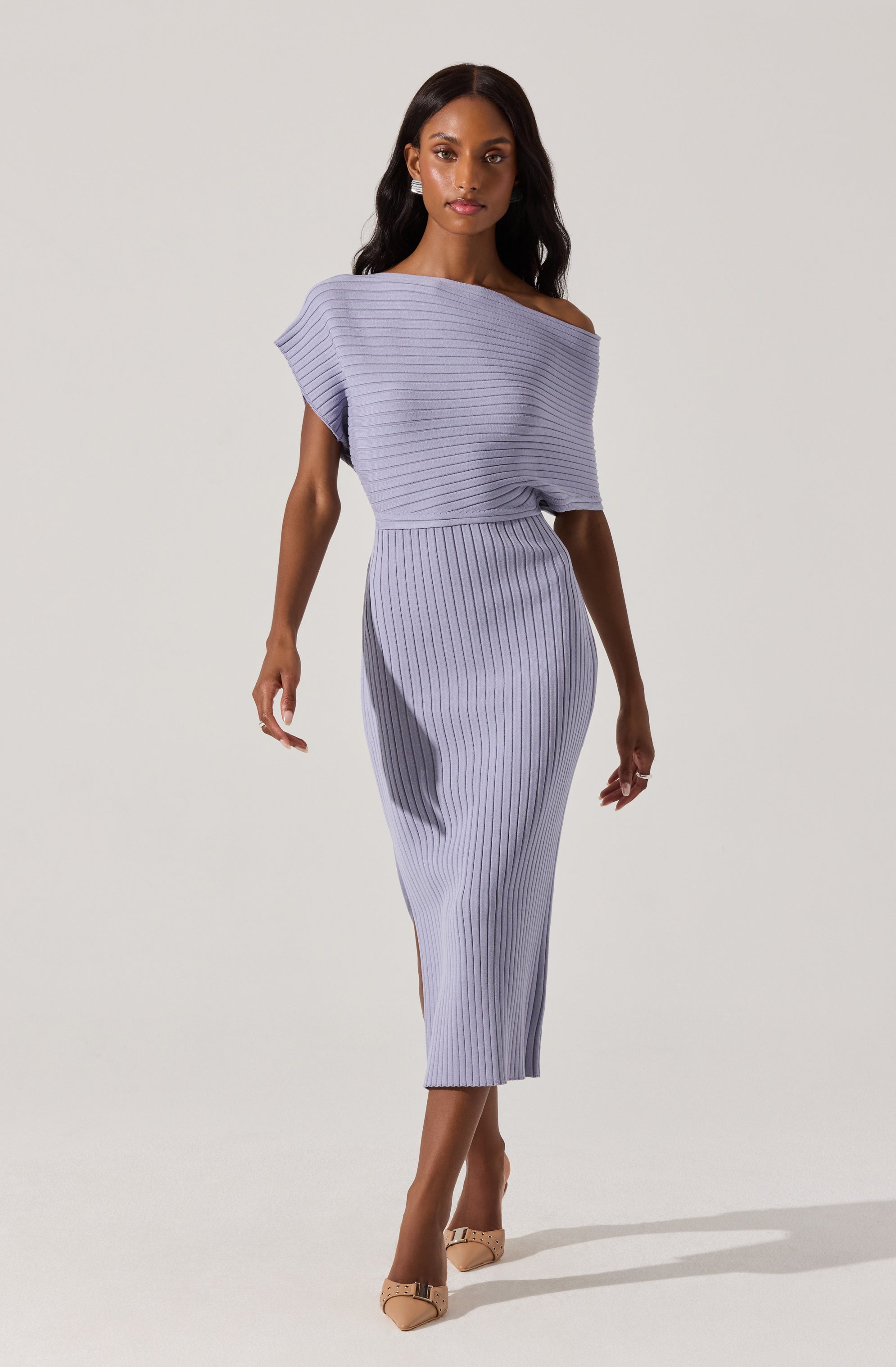 A woman is wearing a light purple, off-the-shoulder dress with thin, vertical striped patterns. The dress has a loose, draped top with short sleeves, and a fitted, high-waisted skirt that extends to mid-calf length. She is also wearing beige high-heeled shoes with decorative embellishments.