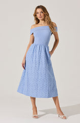 A woman is wearing an off-the-shoulder light blue dress with a fitted, ribbed bodice and a full, midi-length skirt featuring eyelet embroidery. She is also wearing white, open-toe high-heeled sandals.