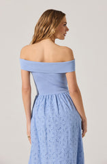 A woman is wearing an off-the-shoulder light blue top with a ribbed texture, fitted at the waist, paired with a long, flowy blue skirt featuring eyelet embroidery and cut-out patterns.