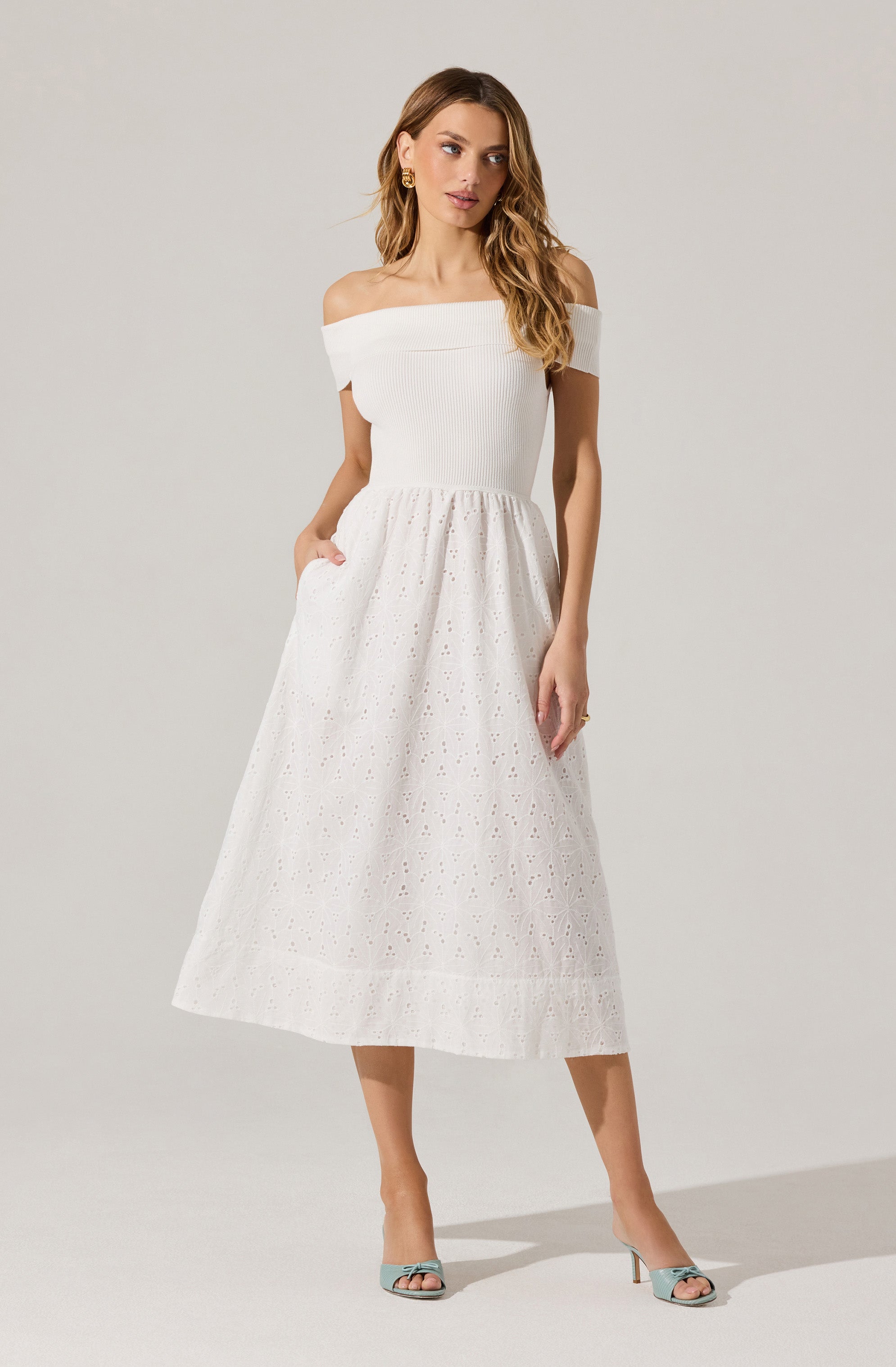 A woman is wearing an off-the-shoulder white dress with a fitted, ribbed bodice and a full, A-line skirt with eyelet embroidery. She is also wearing light-colored high-heeled shoes with an open toe design.