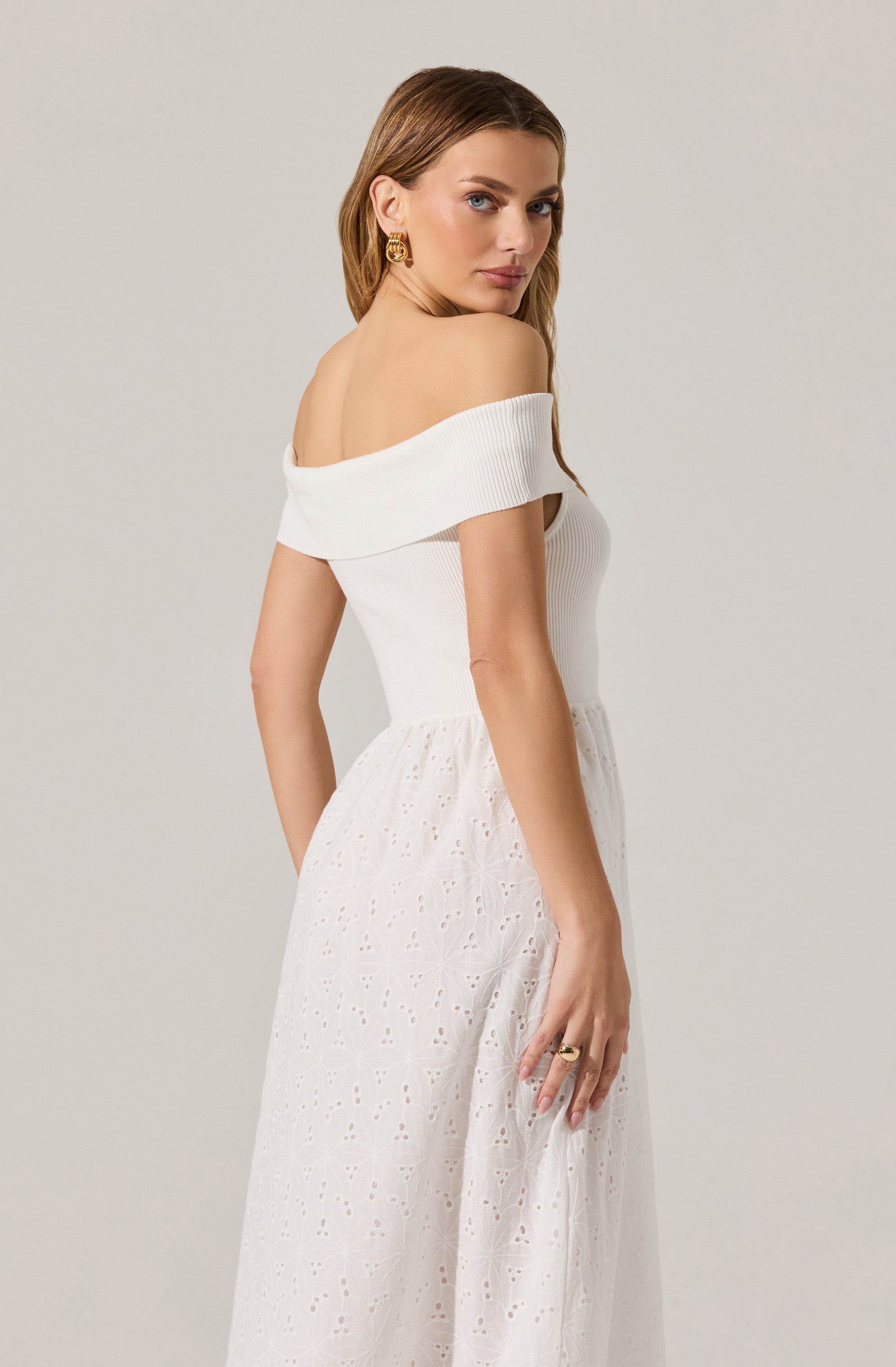A woman is wearing an off-the-shoulder white top with a ribbed texture and a wide fold-over neckline. The top is fitted and short-sleeved, revealing her shoulders and upper arms. She pairs it with a long, white, eyelet-embroidered skirt that has a flowing, airy appearance. She accessorizes with gold hoop earrings and a gold ring on her finger.