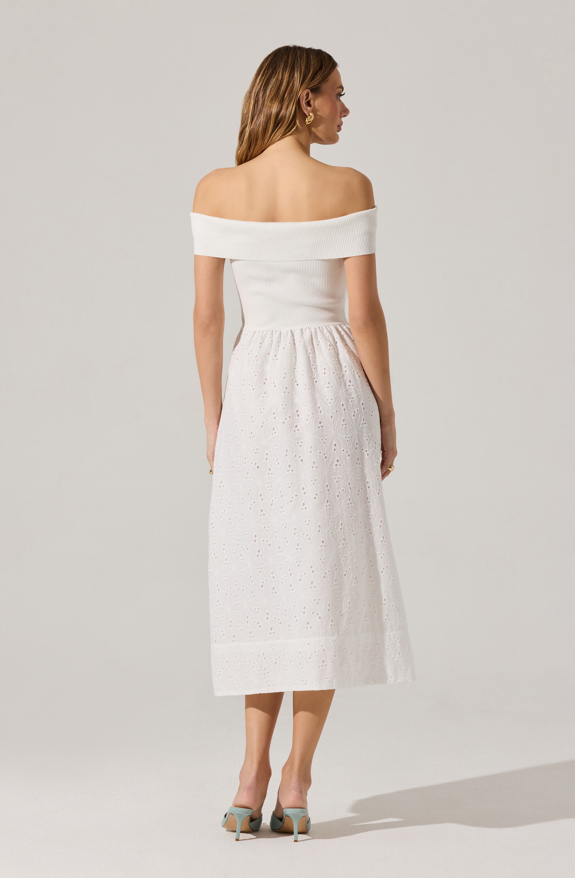 A woman is wearing an off-the-shoulder white knit top with a wide, folded neckline that sits around her shoulders. She is also wearing a white midi skirt made of eyelet fabric with a gathered waist and an intricate, embroidered floral pattern running throughout. She has paired the outfit with light-colored high-heeled shoes.