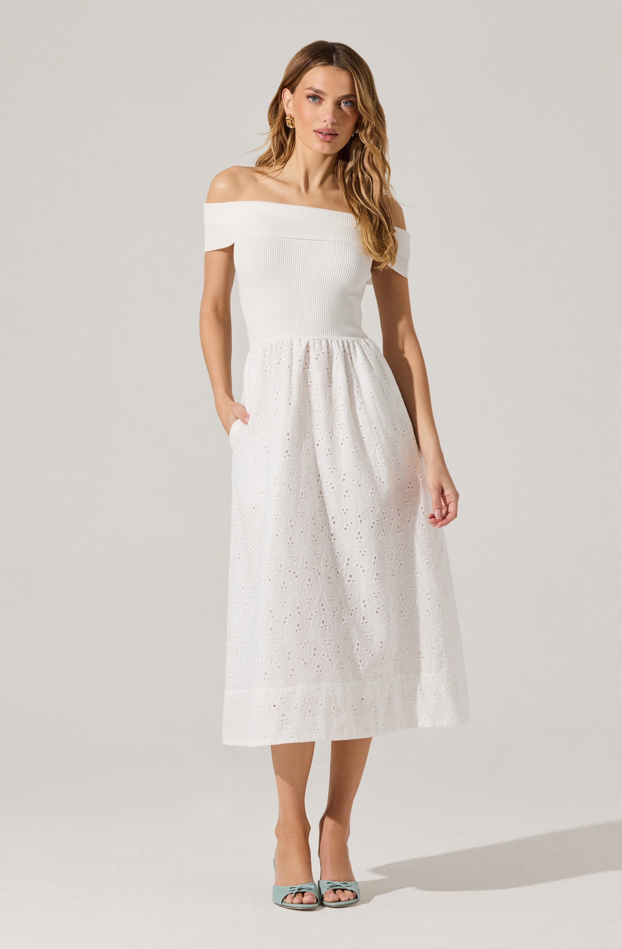 A woman is wearing a white off-the-shoulder dress with a fitted ribbed bodice and a full, eyelet embroidery skirt that reaches mid-calf. She is also wearing open-toed high heels in a light blue color and has gold hoop earrings.