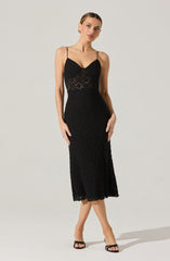 A woman is wearing a black lace midi dress with spaghetti straps, featuring a fitted bodice with a sweetheart neckline and a sheer lace panel at the waist. The skirt falls below the knees with a scalloped hem, and she pairs the dress with black open-toe heels.