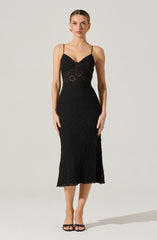A woman is wearing a black, fitted lace dress with thin spaghetti straps. The dress features a sweetheart neckline and the lace pattern is floral and intricate throughout. The hemline of the dress falls to mid-calf length. She is also wearing black open-toed high heels.