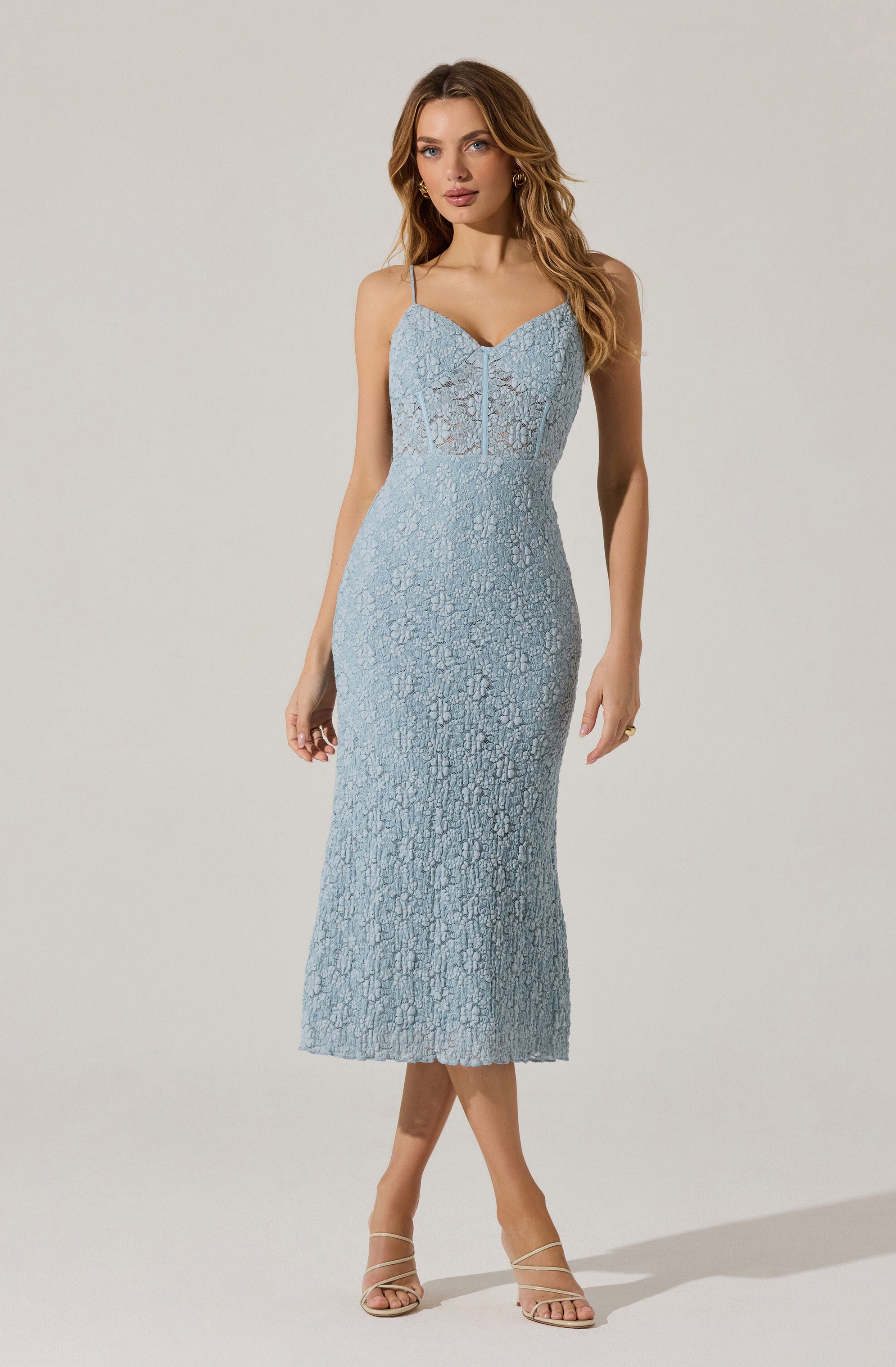 A woman is wearing a fitted, light blue lace dress that features thin spaghetti straps, a sweetheart neckline, and a mid-calf length hem. The dress has intricate lace detailing throughout. She is paired with nude, strappy high-heeled sandals.