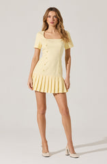 A woman is wearing a light yellow, short-sleeved dress with a square neckline. The dress features a row of buttons down the front and a pleated skirt that extends to mid-thigh. She is also wearing light-colored, high-heeled shoes.