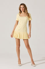 A woman is wearing a short, light yellow dress with a square neckline and short sleeves. The dress features a row of buttons down the front and a pleated skirt that extends slightly below the waist. She is also wearing light-colored heels.