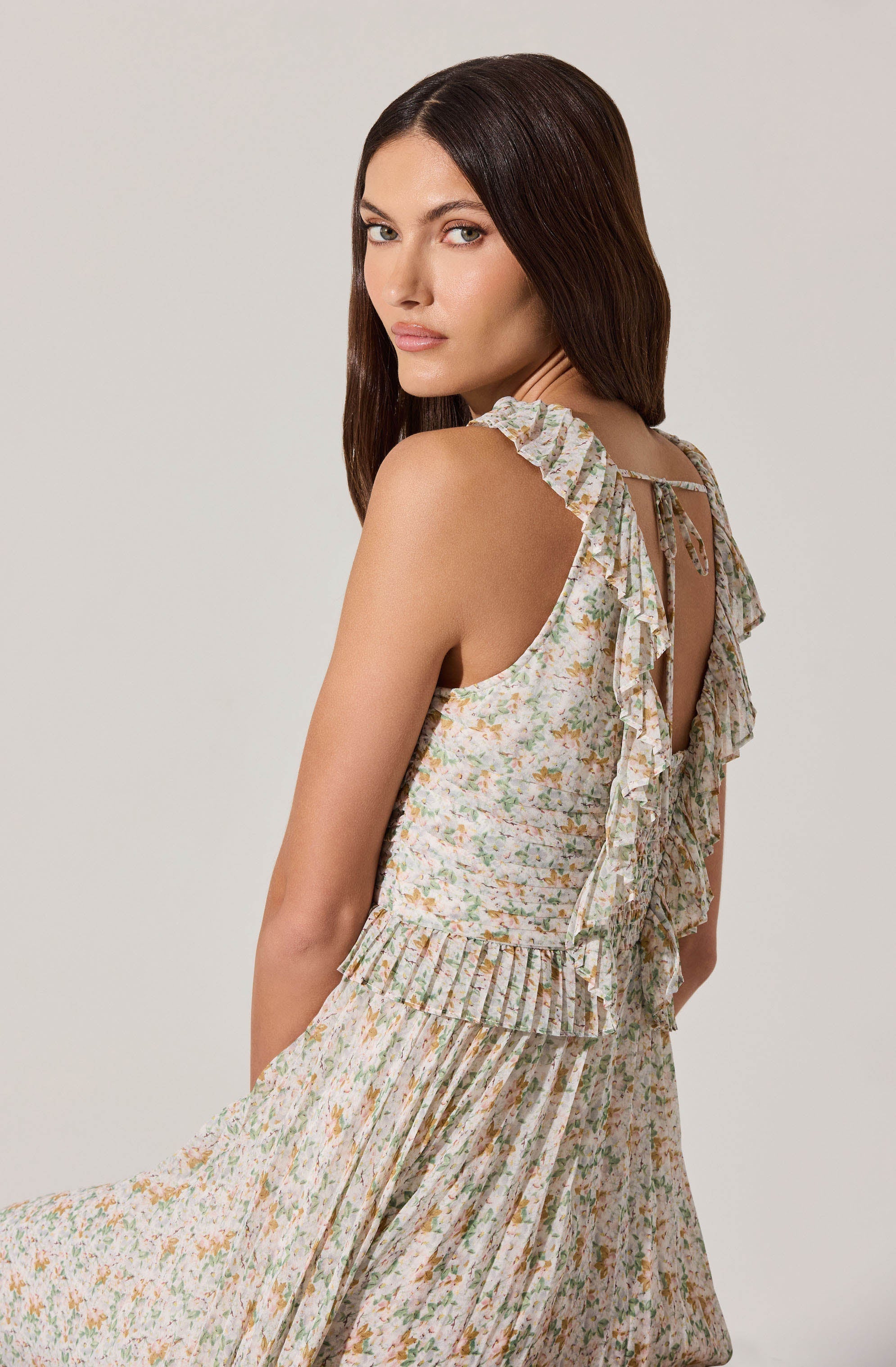 A woman is wearing a sleeveless, floral-patterned dress with ruffled details along the shoulder straps and neck. The dress features a light, pastel-colored print with a mix of green, peach, and beige tones. The fabric appears lightweight, and the dress has a fitted bodice with a gathered and pleated skirt. The overall look is feminine and summery.