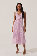 A woman is wearing a sleeveless, V-neck dress with a fitted bodice and a pleated skirt. The dress features a pastel pink and lavender floral pattern, with a ruffle detail at the neckline and a fabric bow at the waist. She is also wearing open-toe high heel sandals in a nude color.