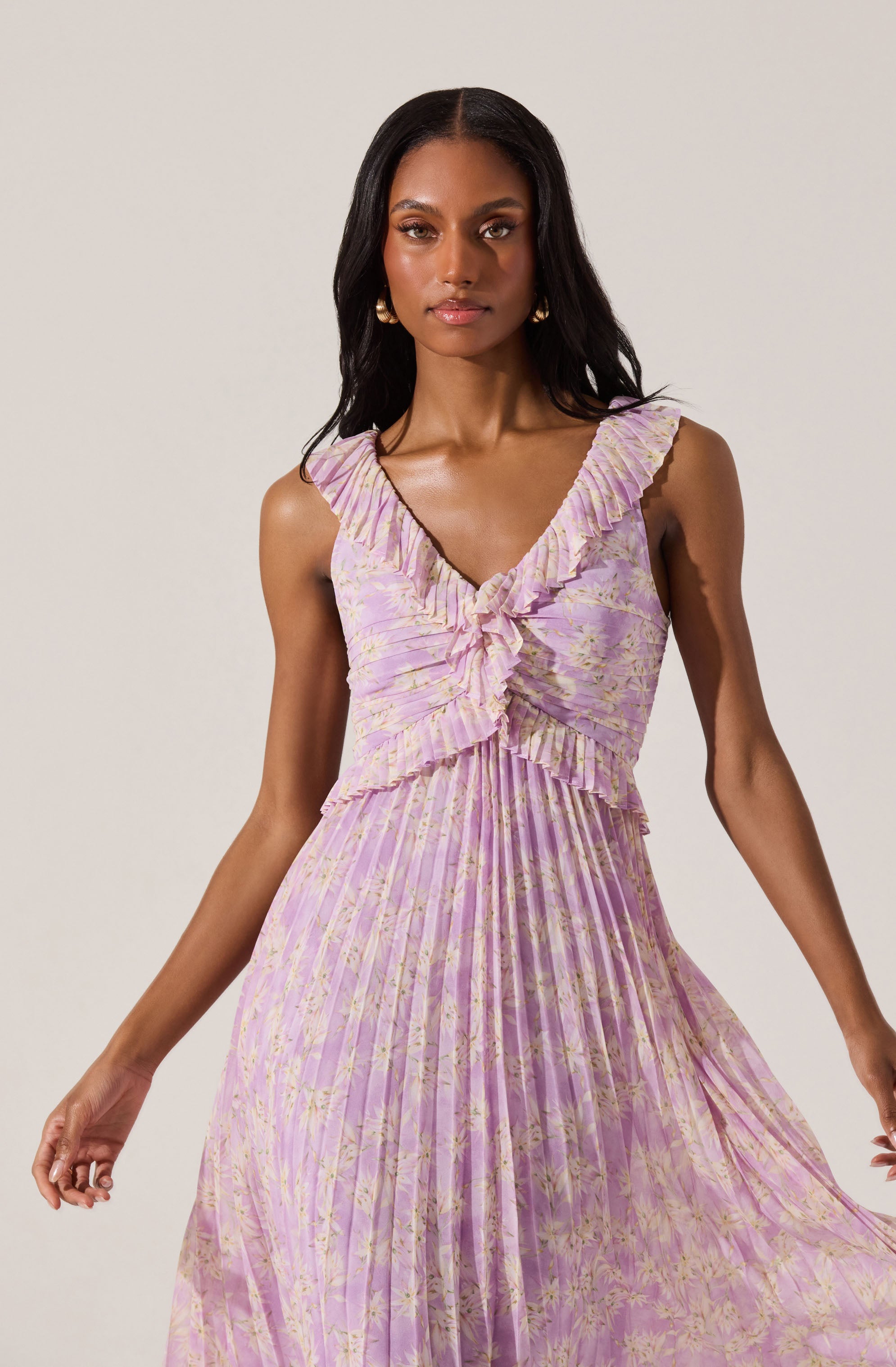 A woman is wearing a sleeveless, ankle-length dress with a V-neckline and ruffled trim. The dress is made of a pleated fabric with a light pink and lavender tie-dye or floral pattern. The bodice features a ruffled detail that cascades from the shoulders down the front.
