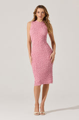 A woman is wearing a sleeveless, knee-length pink textured dress with a high neckline. She is also wearing nude-colored open-toe heels and gold earrings.