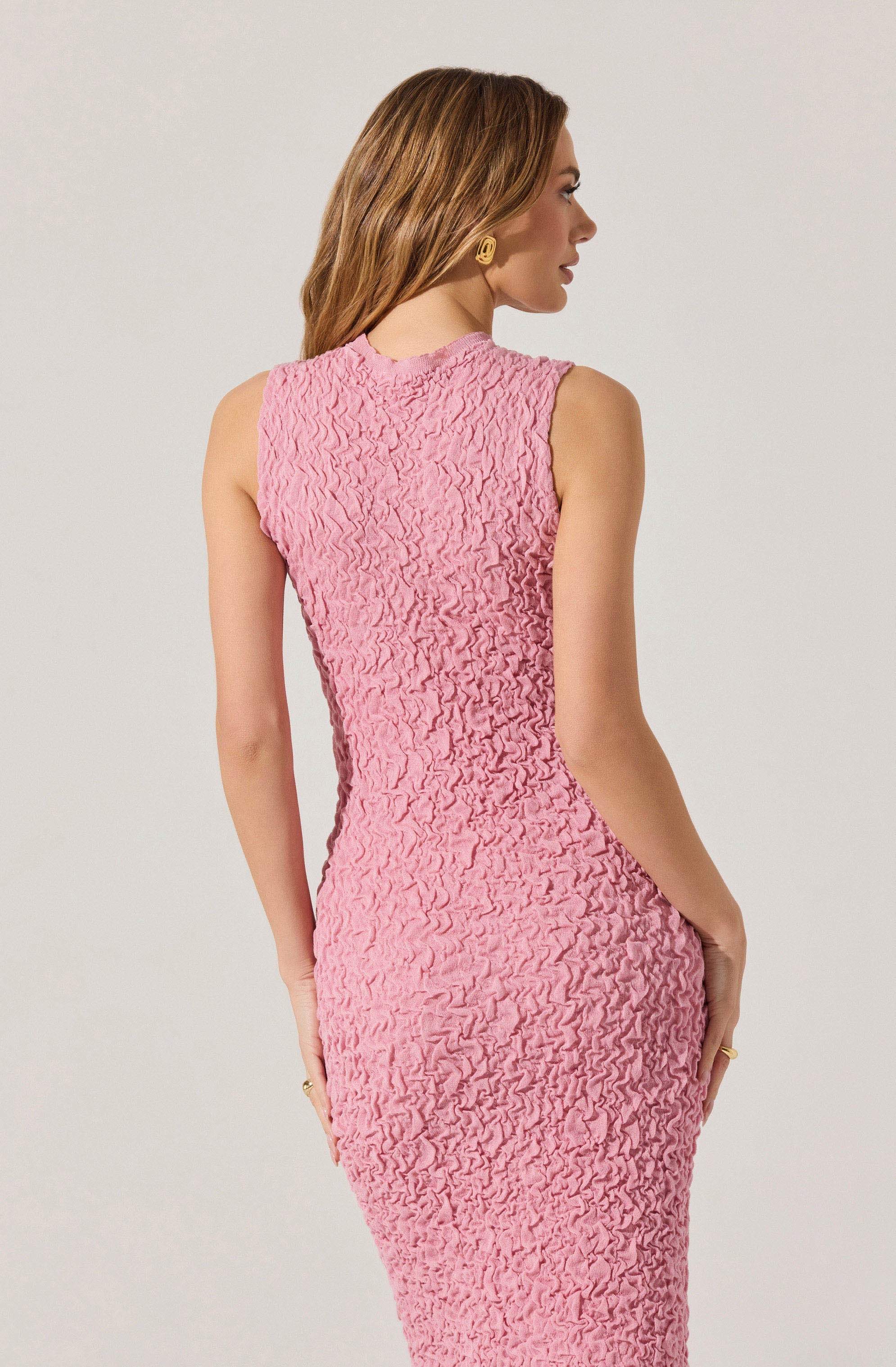 A woman is wearing a sleeveless, fitted dress in a soft pink color with a textured, ruched fabric. The dress has a high neckline and is form-fitting, accentuating her waist and hips.