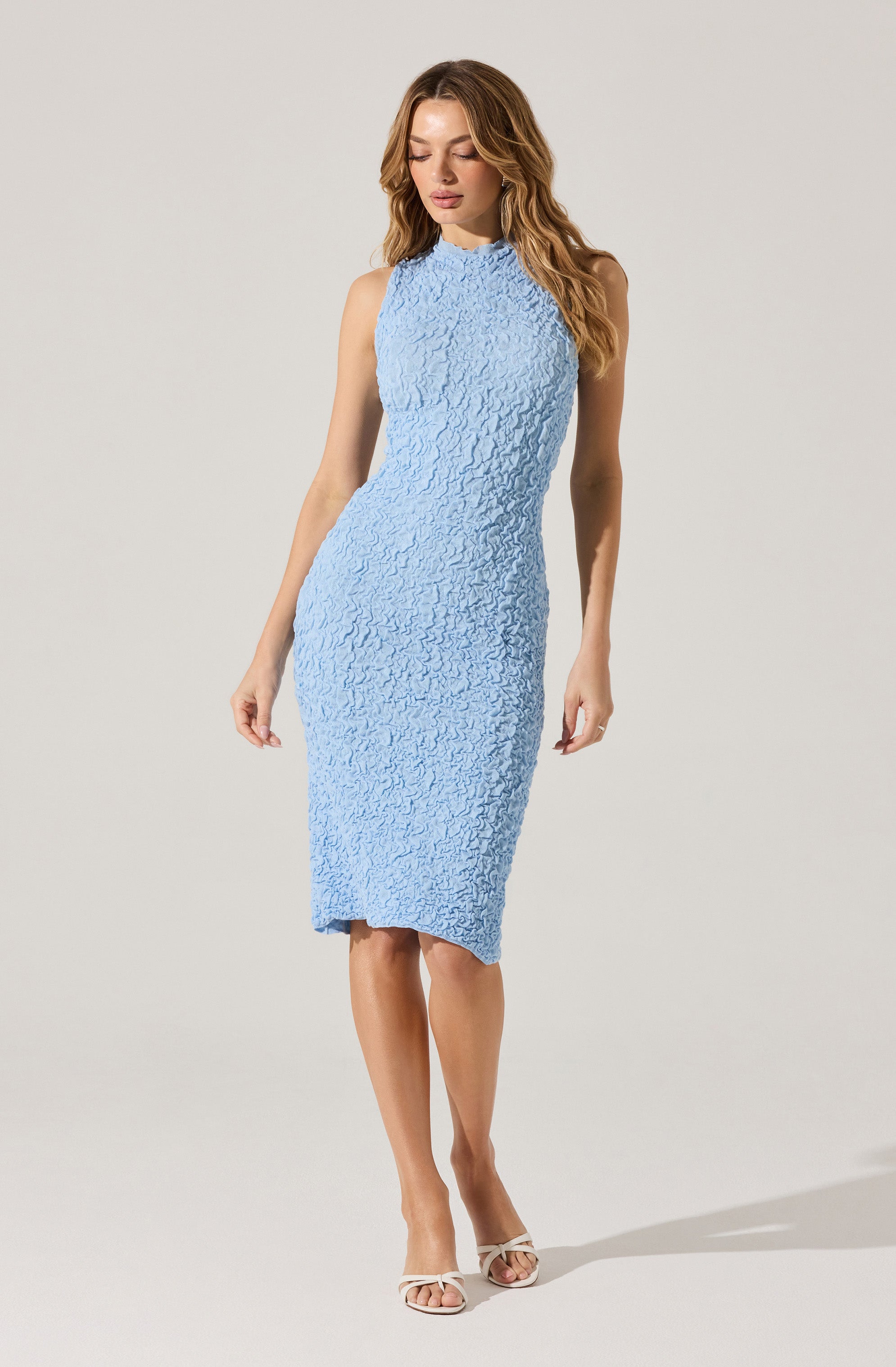 A woman is wearing a sleeveless, knee-length dress in a light blue color with a textured, crinkled fabric. She has on white open-toe heels.