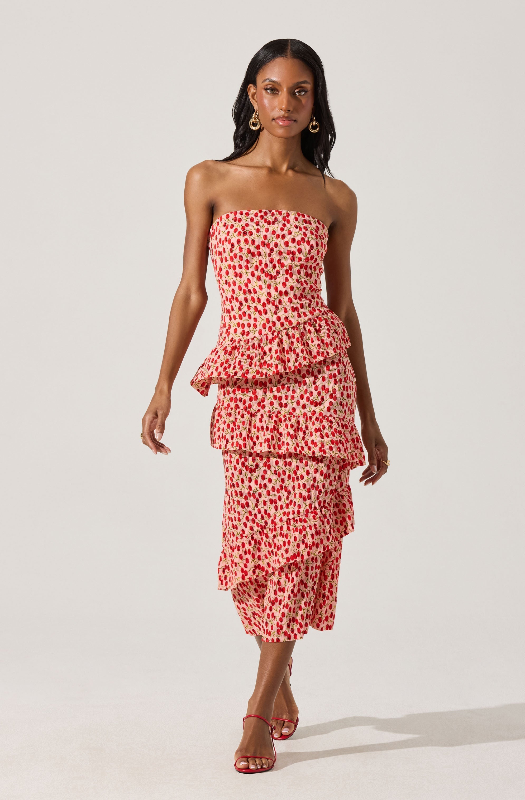 A woman is wearing a strapless, off-the-shoulder dress with a beige background and a red and pink dotted pattern. The dress features three ruffled tiers, creating a layered look. She is also wearing red strappy high-heeled sandals and gold hoop earrings.