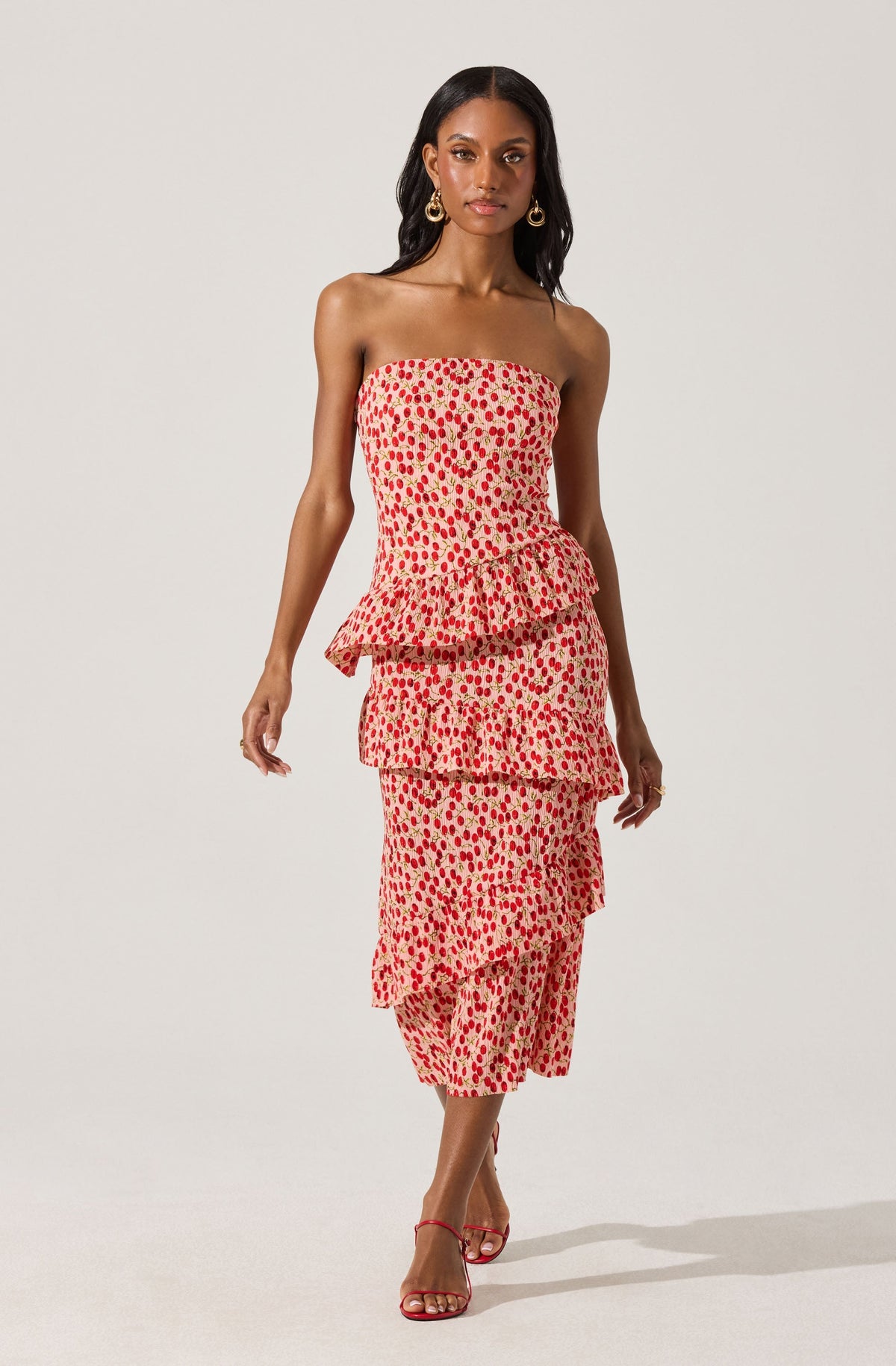 A woman is wearing a strapless, off-the-shoulder dress with a beige background and a red and pink dotted pattern. The dress features three ruffled tiers, creating a layered look. She is also wearing red strappy high-heeled sandals and gold hoop earrings.