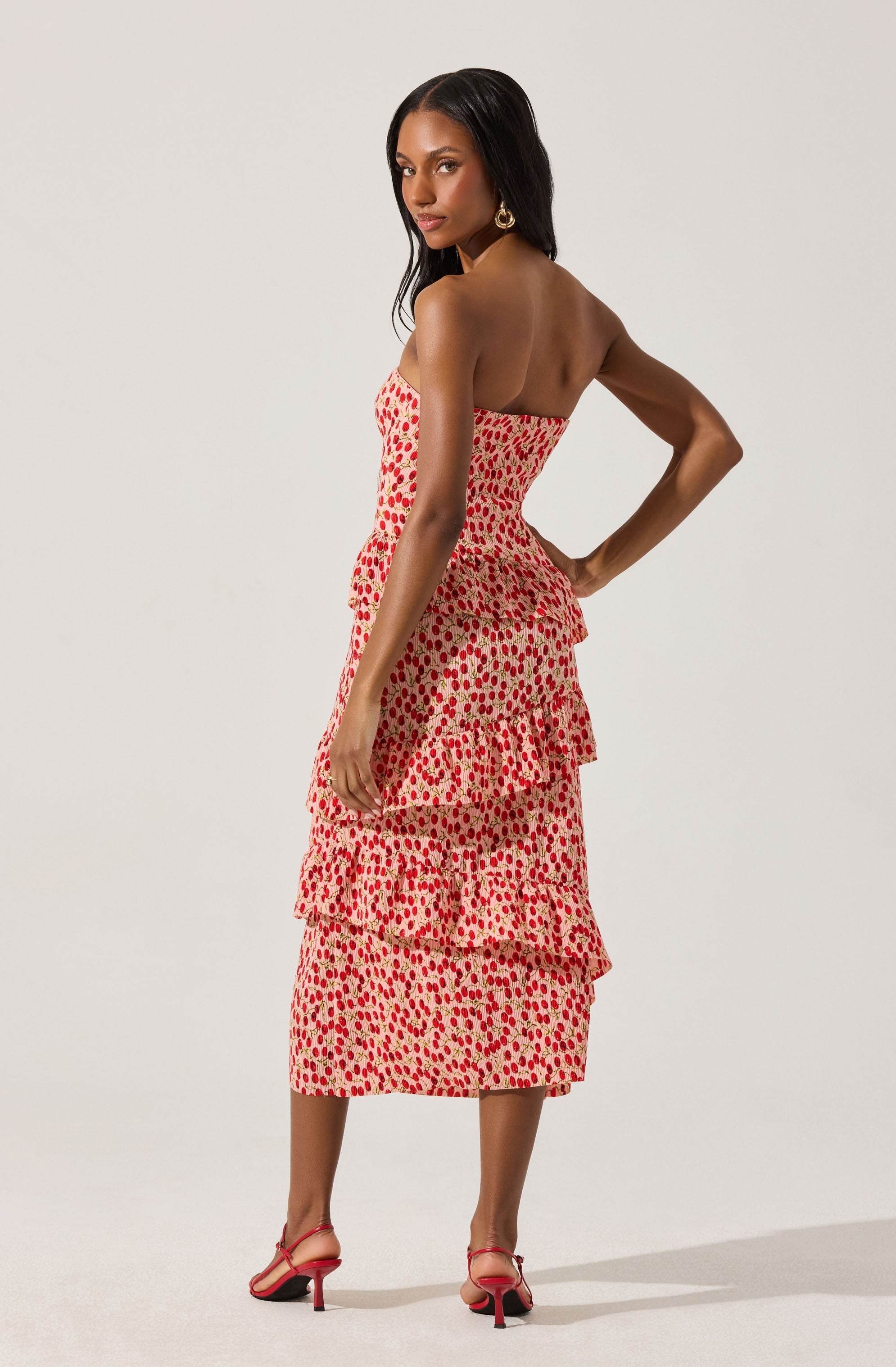 A woman is wearing a strapless, knee-length dress with a beige background and red, pink, and orange polka dot pattern. The dress features a fitted bodice and three layered ruffles on the skirt. She is also wearing pink high-heeled sandals with ankle straps and large gold hoop earrings.